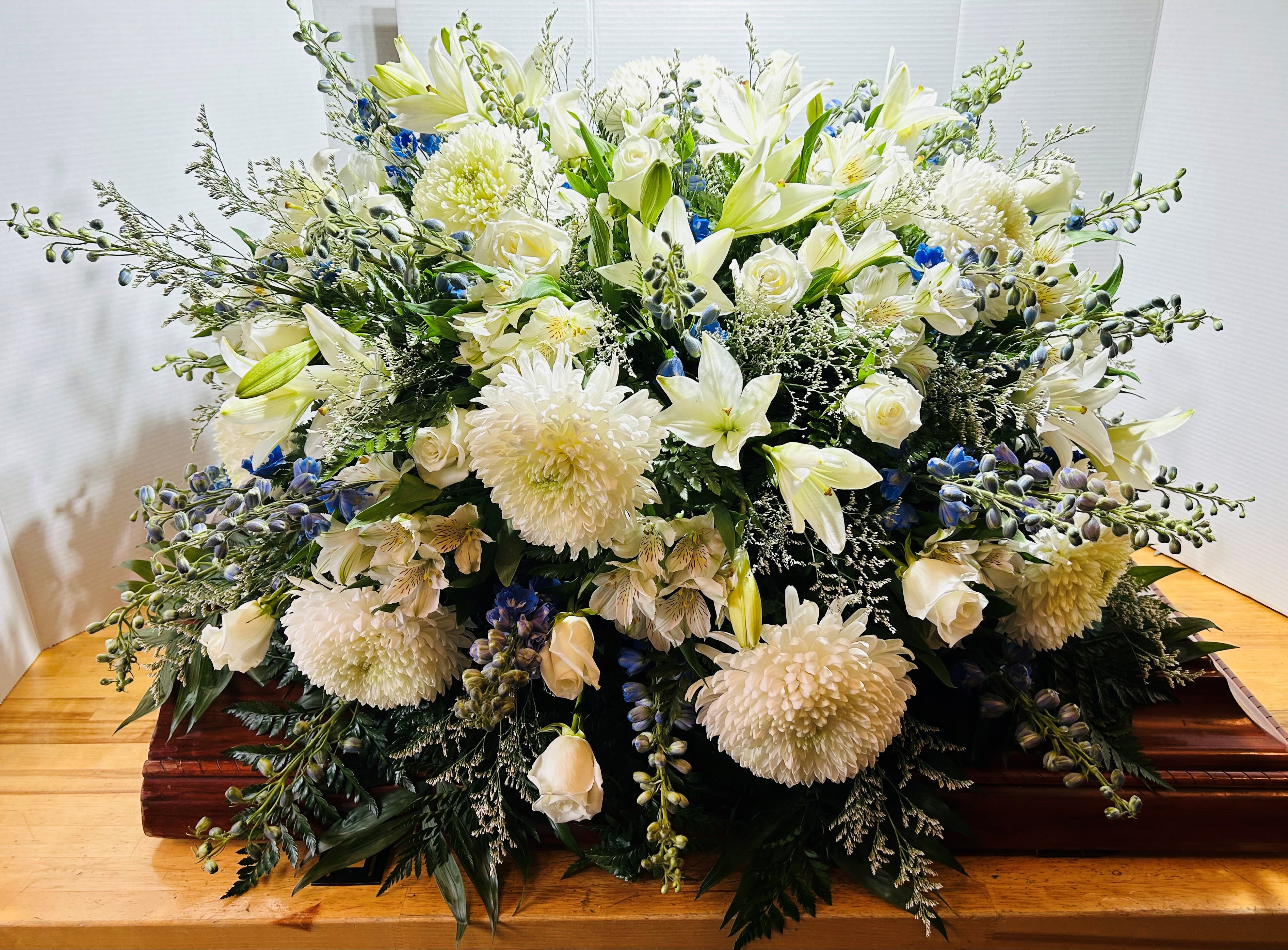 Bountiful Memories Casket Spray Bountiful Memories Casket Spray - Beautiful blue and white casket spray is delivered decorated with white mums, white roses, blue delphinium, white alstroemeria, white lily's, and Limonium along with emerald palm and leather leaf.