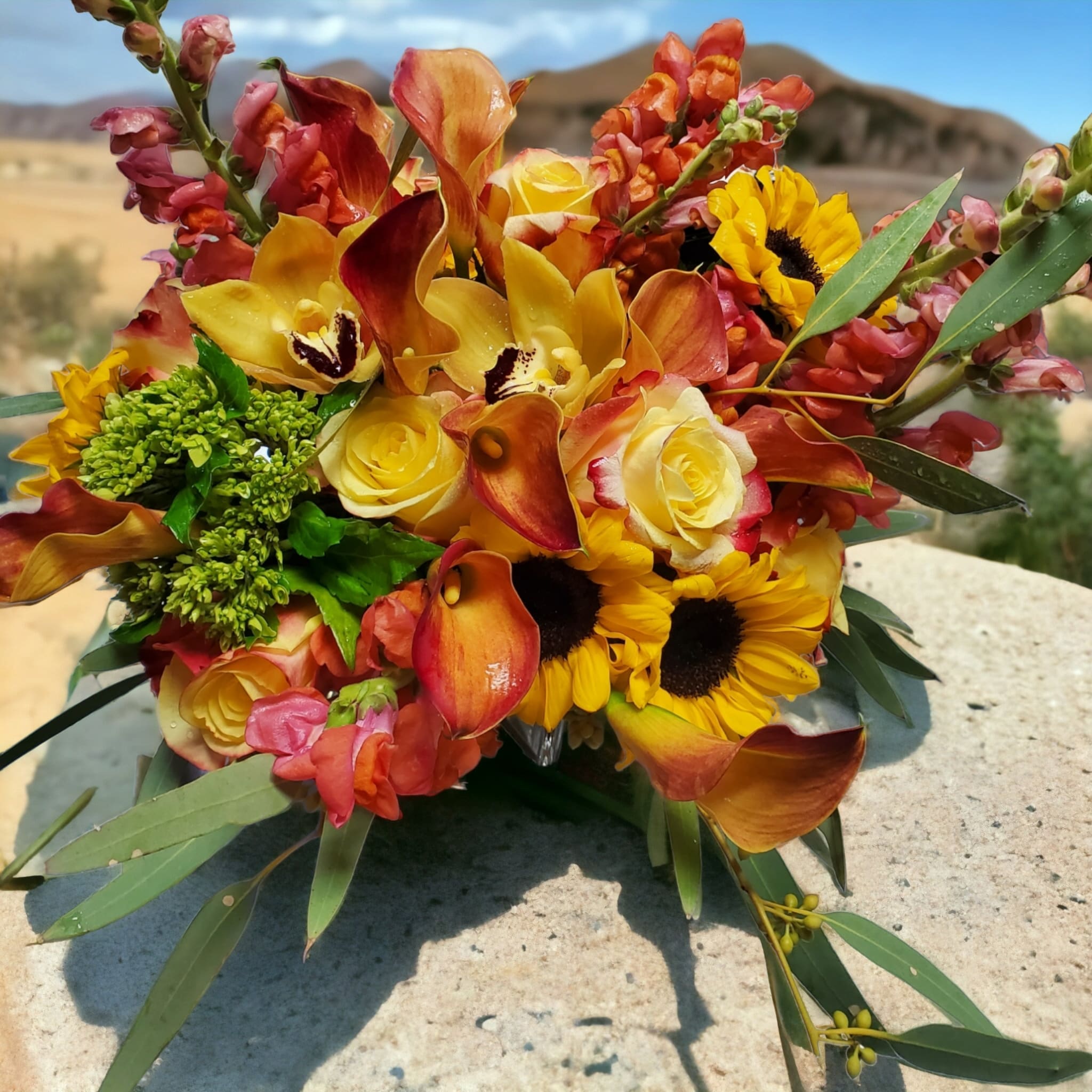 Arizona Sunrise - Rich golden yellows, orange and shades of burgundy featuring Callas, Sunflowers, Roses, Cymbidium Orchids and other seasonal flowers creatively designed in a crystal clear cube.  Substitutions may be needed based on availability of flowers,  but the general feeling will remain the same.