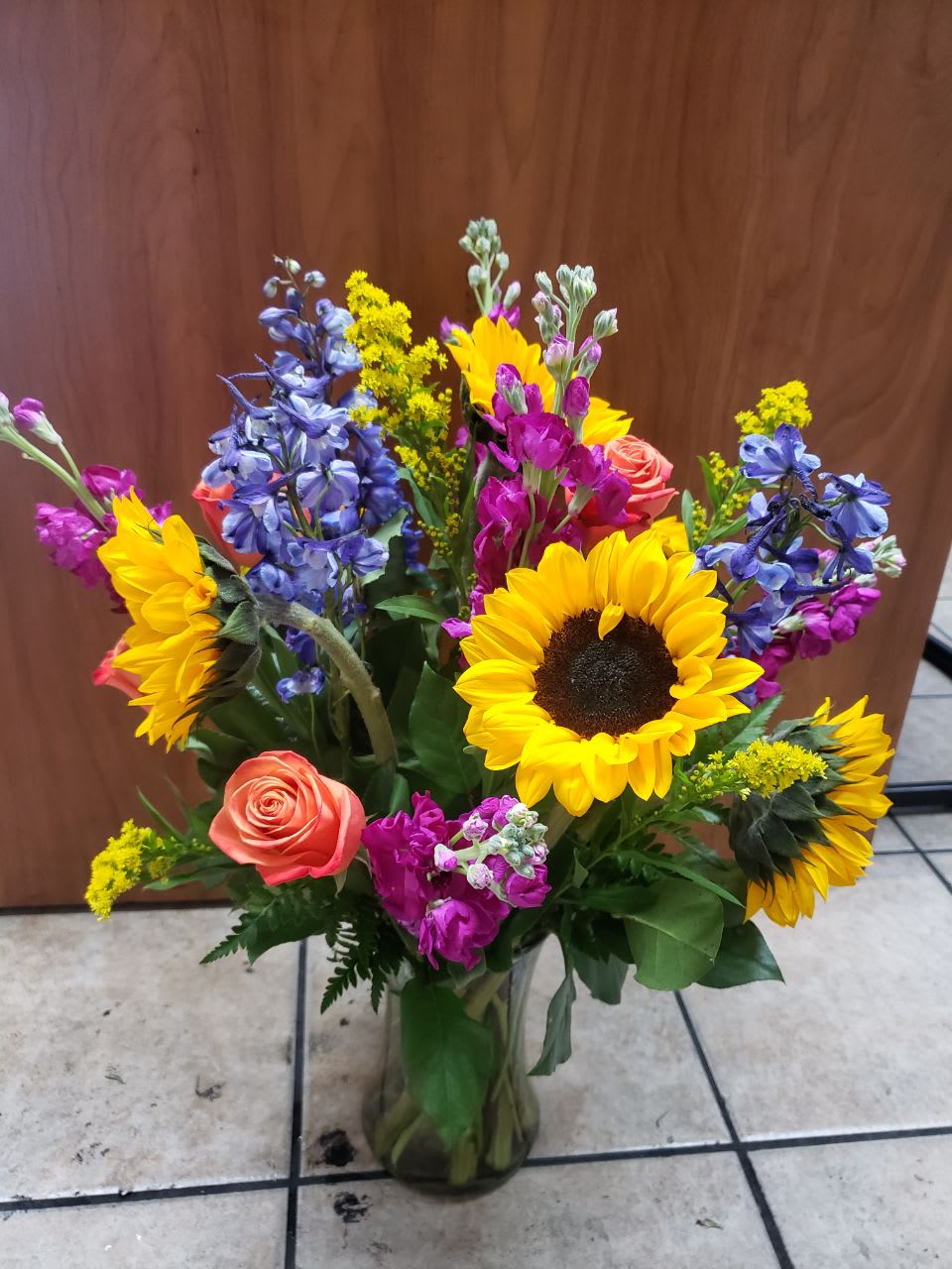 Rainbow Bits - This beautiful arrangement, you can feel the rainbow in your hands,  has  6 medium sunflowers,  5 oranges roses,5  fuchsia  stock , 5 stems of blue delphinium and 3 stems of yellow solidago, salad tips and leatherleaf stems. in a medium  clear gathering  vase,