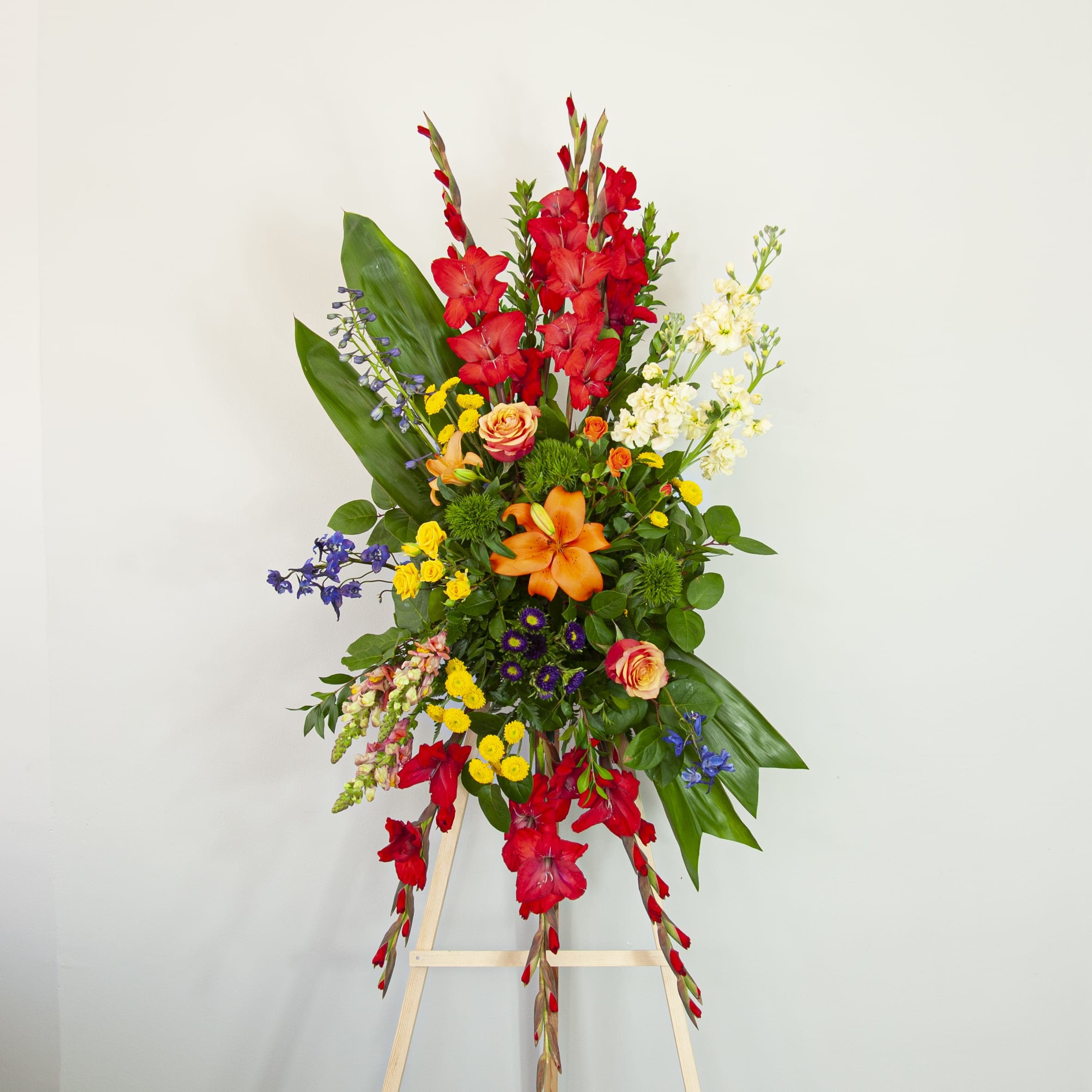 Sunset Memories - Standing Spray (EXP 46) - This colorful standing spray is made with a sunset-inspired color palette, and will brighten up the mood to this somber occasion. Let it be a memorable tribute to one who was loved so dearly, our hearts go out to your time of loss and mourning.   Blooms: - red gladiolas - orange lilies - white stock - blue delphinium - orange and yellow garden roses - yellow and green button poms 