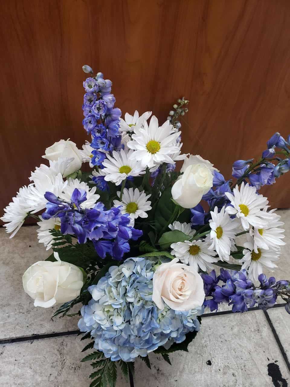 Feel The Sky - A perfect combination of blue like the sky and white like the clouds that make this arrangement unique like our sky 4 stems of white daisies, 2 blue hydrangeas, 4 white roses, 5 stems of blue delphiniums, salad tips, fern leaf in a small cinched vase
