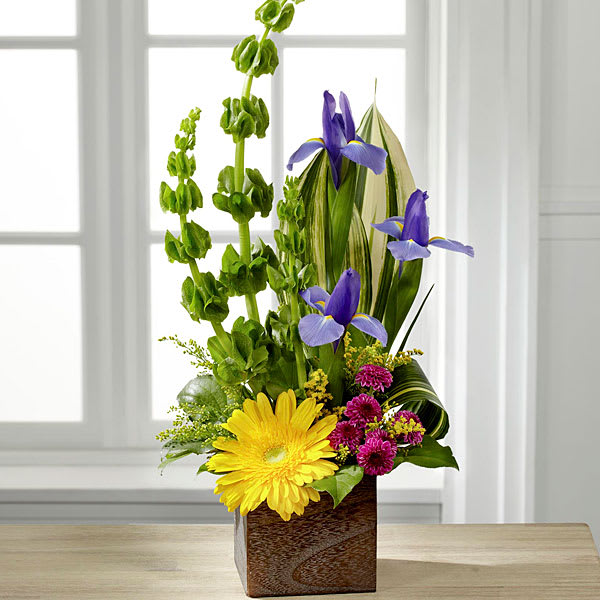 Best Year Arrangement - Whether for a birthday in celebration of the New Year or even as a way congratulate your recipient on a new job or adventure this bouquet conveys your warmest wishes with each sunlit bloom. Happy and bright gerbera daisies are at the base of this arrangement with brilliant green stalks of Bells of Ireland blue iris and tropical leaves stretching upwards for a vertical display. Accented with yellow solidago purple button poms and lush greens this stunning flower arrangement arrives in a rustic square wooden planter to give it a natural background in which to truly shine. 