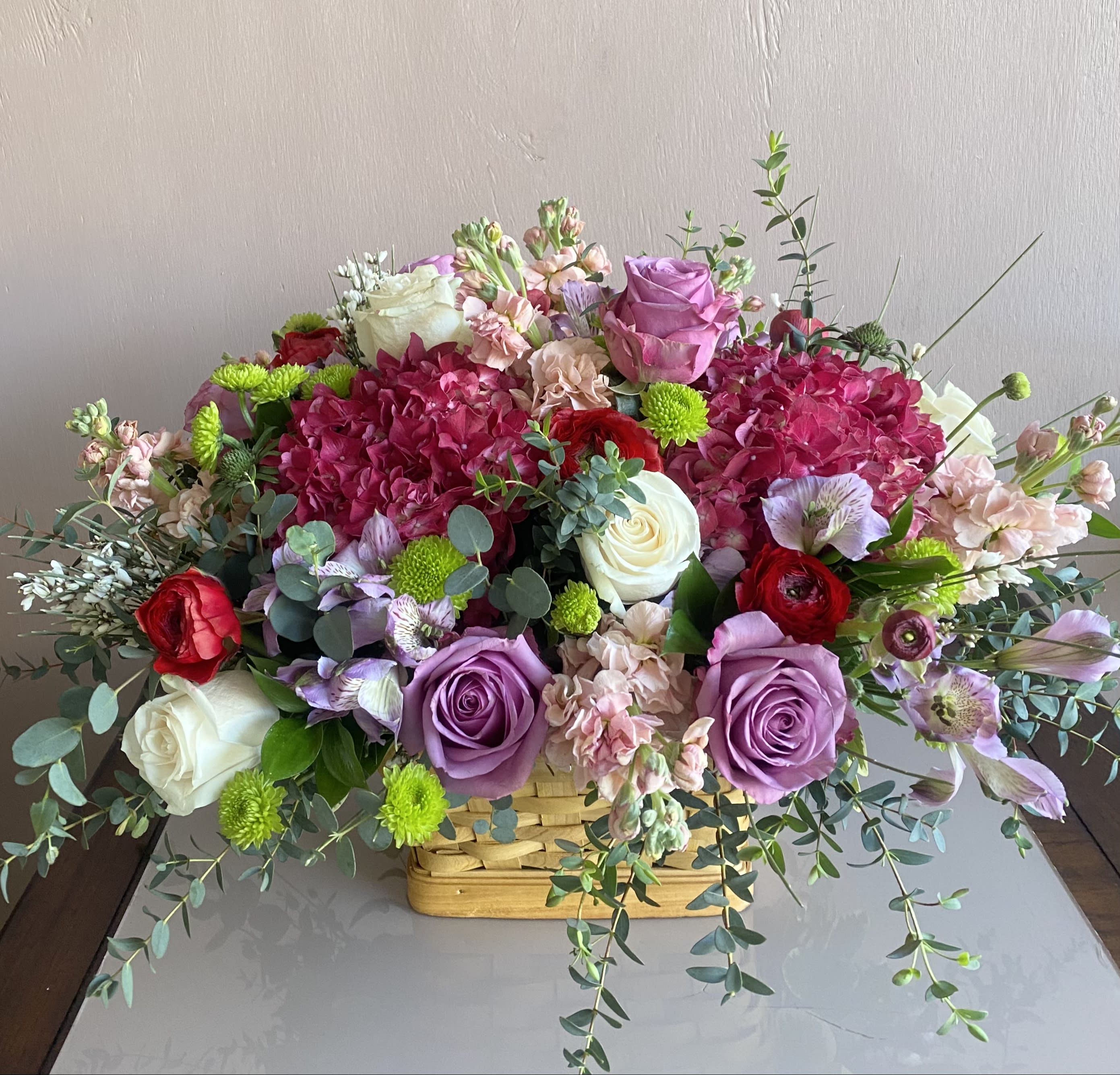 Colorful Basket - An arrangement includes hydrangeas, roses, ranunculus, stock, alstroemeria, and foliage in a wooden basket standing 12&quot; tall x 16&quot; wide. Please note base on the market, maybe the designer will use a substitute for the flowers. 