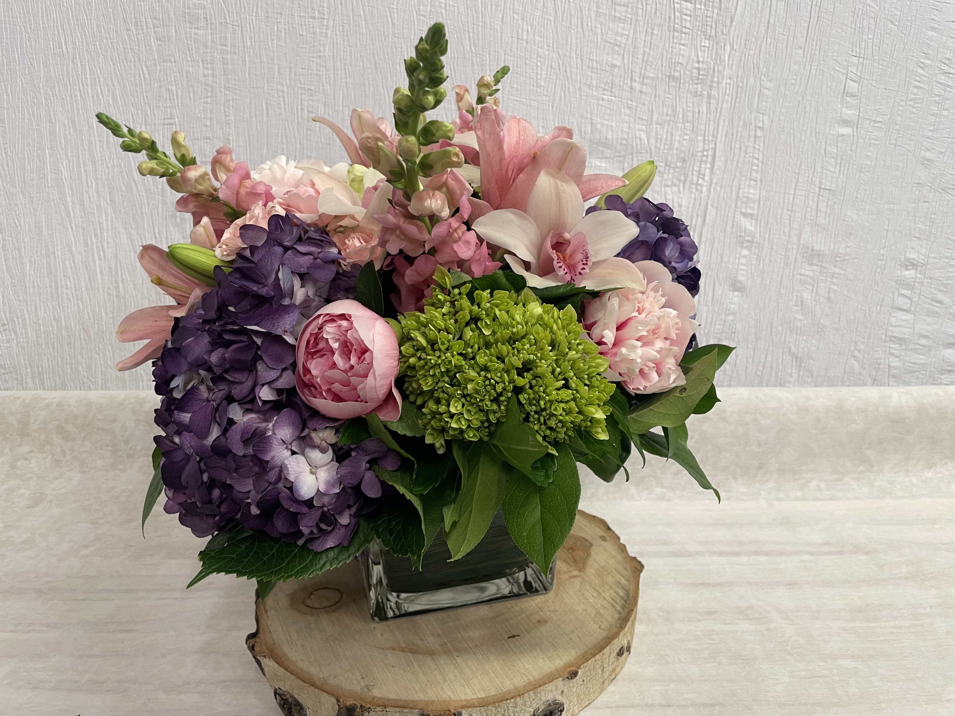 Sweet Song Arrangement  - This stunning arrangement is made in a leaf lined glass cube making it very modern and sophisticated. The assortment of deep purple hydrangea paired with bright green and pink assorted flowers gives this arrangement a sweet style. 