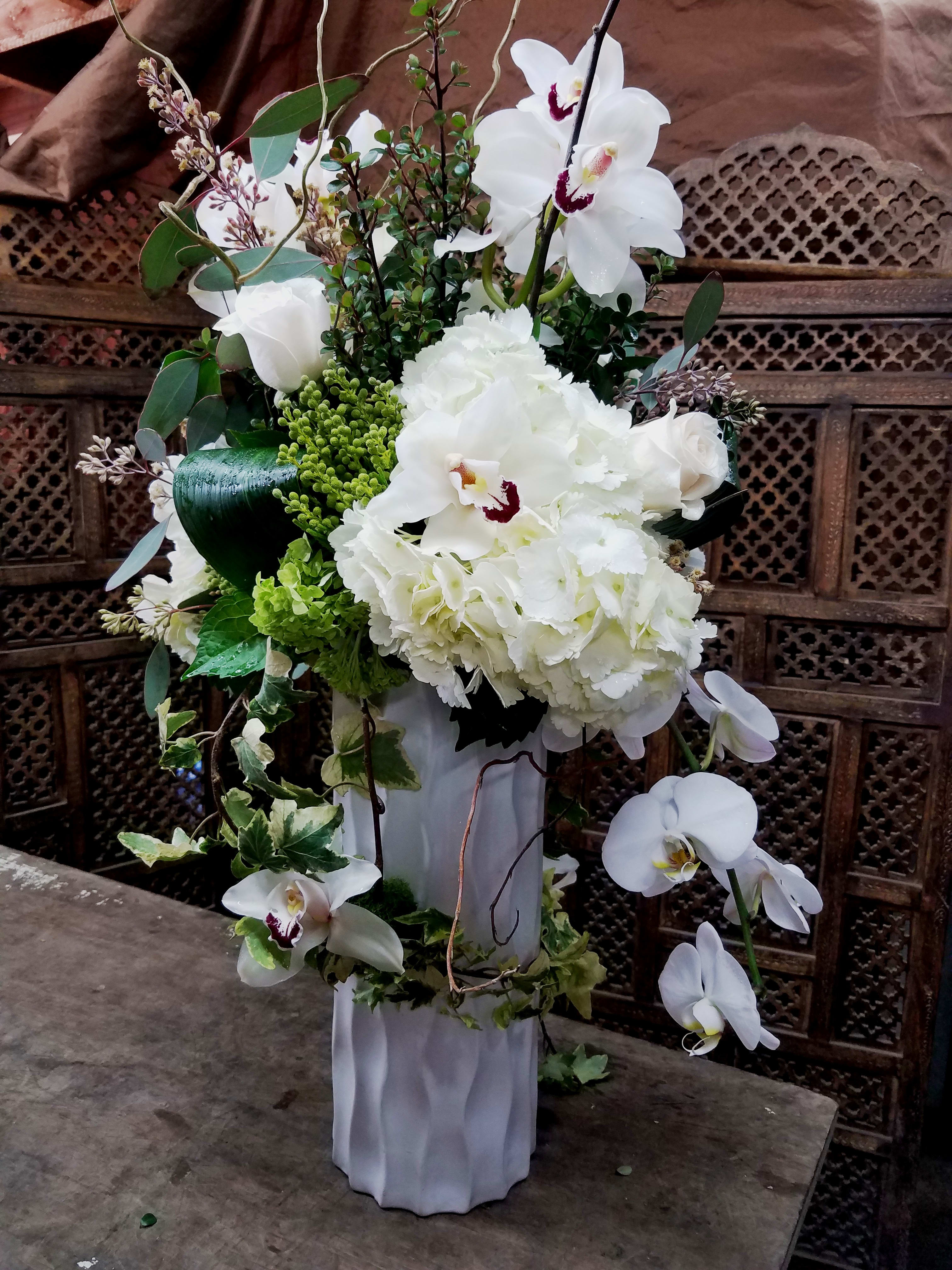 Topsy-Turvy in White - A taller arrangement which juxtaposes Cascading White Phaelenopsis Orchids with Rising Cymbidium Orchids and an upshot of Branches and White Roses.  This design also has a playful swoosh of Variegated Ivy.  it really has that &quot;Wow&quot; factor of beauty and is even more stunning in Premium version.
