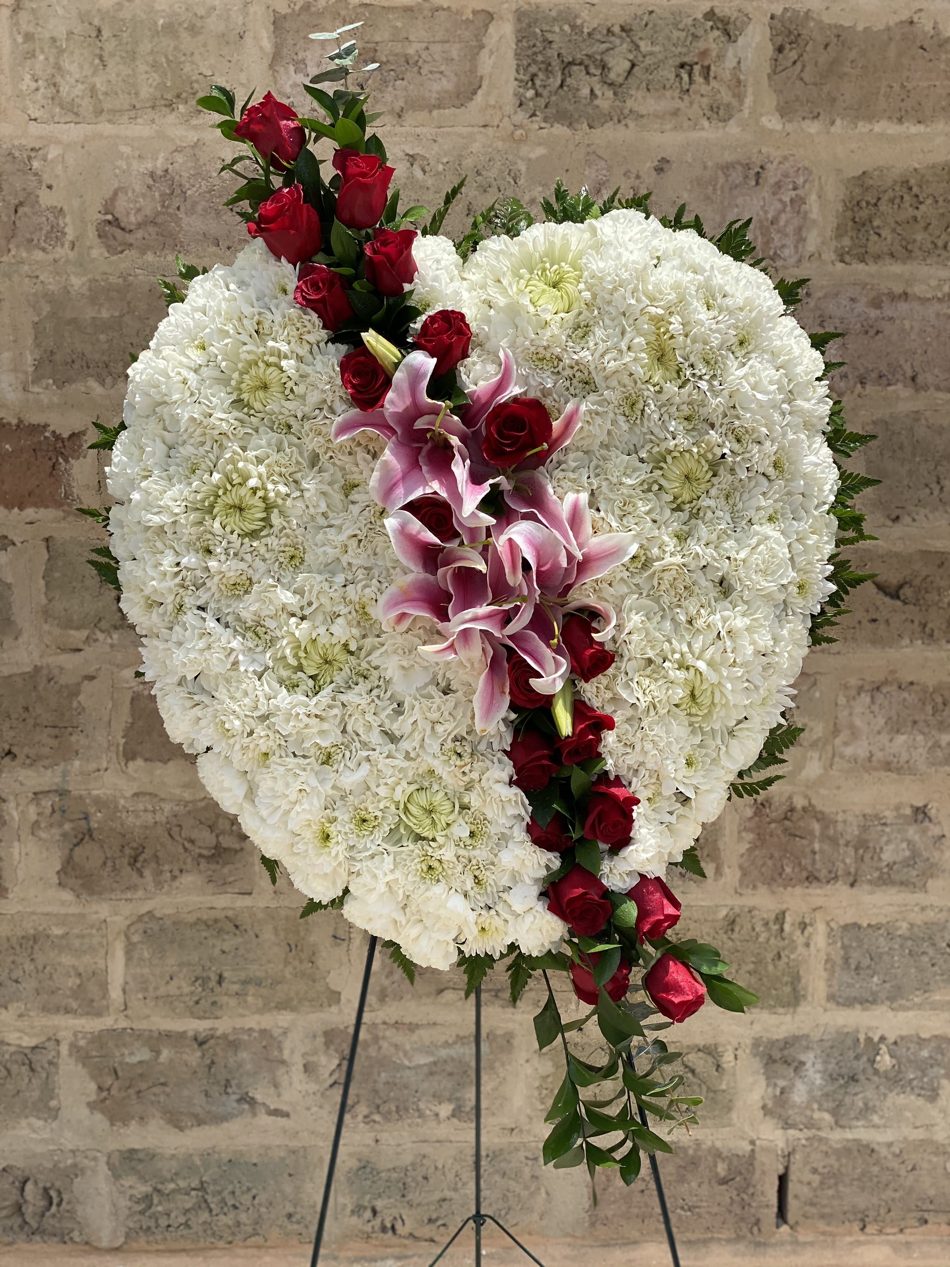 Majestic Heart - This beautiful floral tribute shows your undying love during this difficult time. This standing spray arrangement in the shape of a heart is created from fresh white flowers with red roses.Traditionally sent directly to the funeral home by family members or friends and displayed on a stand. Our florists use only the freshest flowers available so varieties and colors may vary.