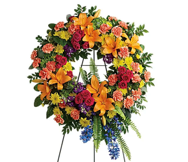 Colorful Serenity Wreath - A rainbow of love. Celebrate your colorful memories of a beautiful life with this radiant wreath of hydrangea roses and lilies. This wreath features green hydrangea hot pink spray roses orange asiatic lilies orange carnations blue delphinium purple stock yellow cushion spray chrysanthemums huckleberry variegated pittosporum sword fern silver dollar eucalyptus and lemon leaf. Delivered on a wire easel.
