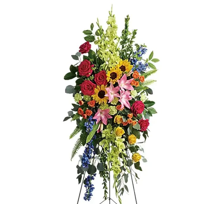 Love Lives On Spray - Bursting with joy this radiant spray of rainbow blooms is an exuberant tribute to never-ending love. This spray features hot pink roses yellow roses orange spray roses pink asiatic lilies green gladioli green carnations medium yellow sunflowers blue delphinium bupleurum myrtle sword fern silver dollar eucalyptus and lemon leaf. Delivered on a wire easel.