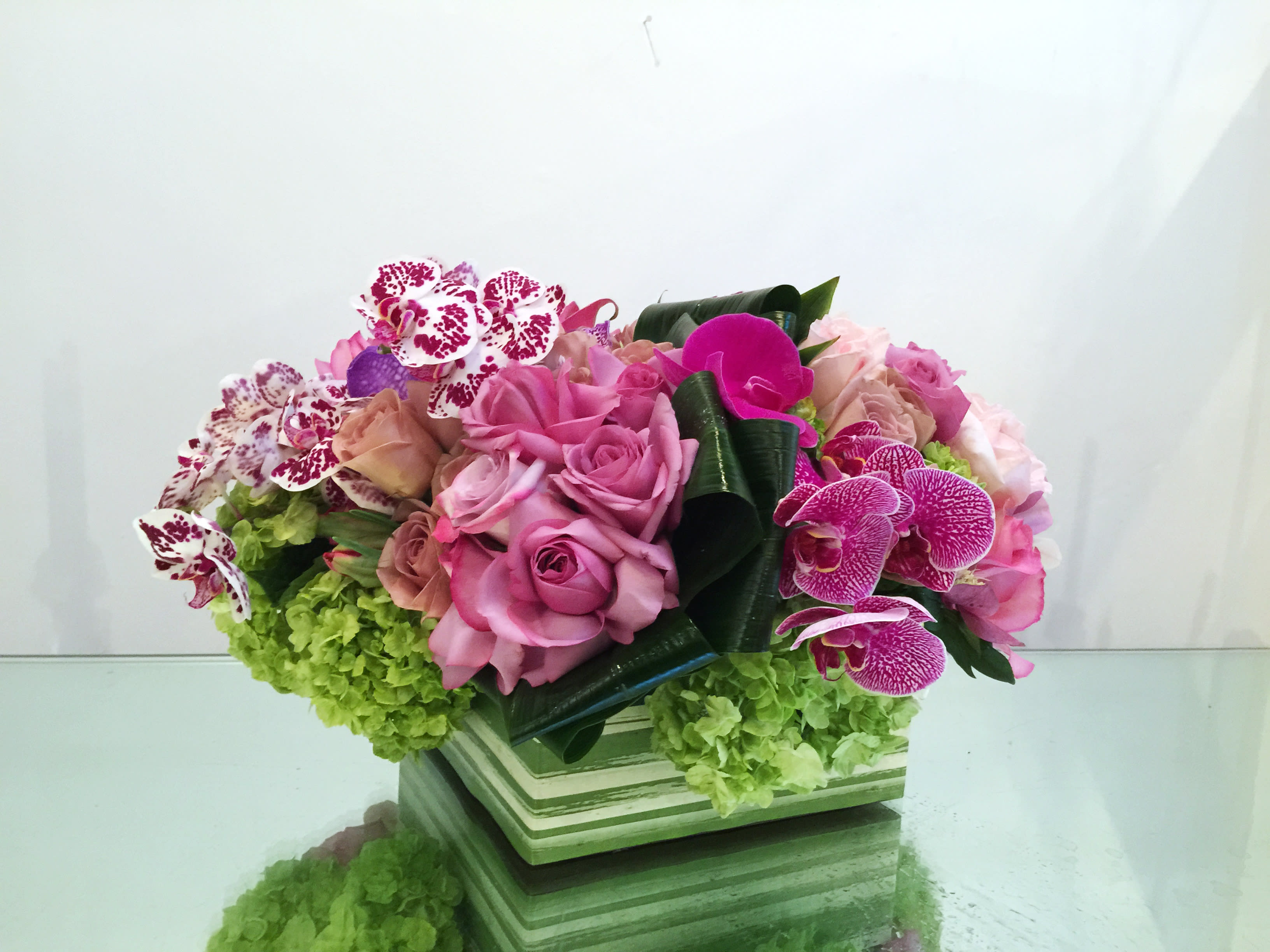 Secret Garden - A luxurious and fresh springtime arrangement with bright green hydrangeas, dusty pink roses, and various purple phalaenopsis orchids with modern green accents in a decorative square glass vase.  *PREMIUM pictured*