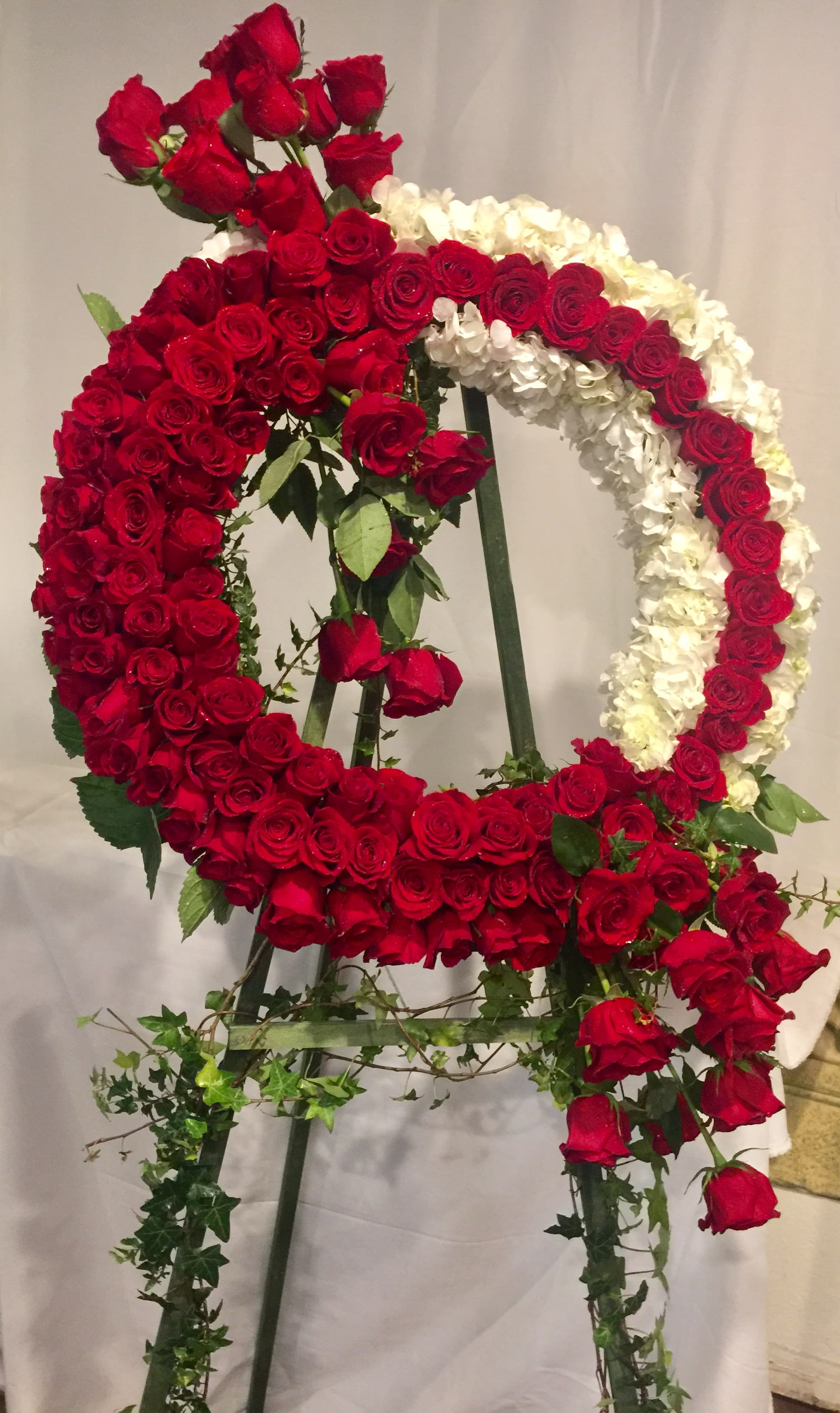 SYM01 - Red and White Wreath - The Red and White Wreath is the perfect way to honor the life of a loved one or add a touch of elegance to a special event. This stunning 24&quot; floral wreath includes a mix of red roses and white hydrangea in a handcrafted, sleek design, accented with flowing greenery. The Red and White Wreath is attached to a sturdy easel. 