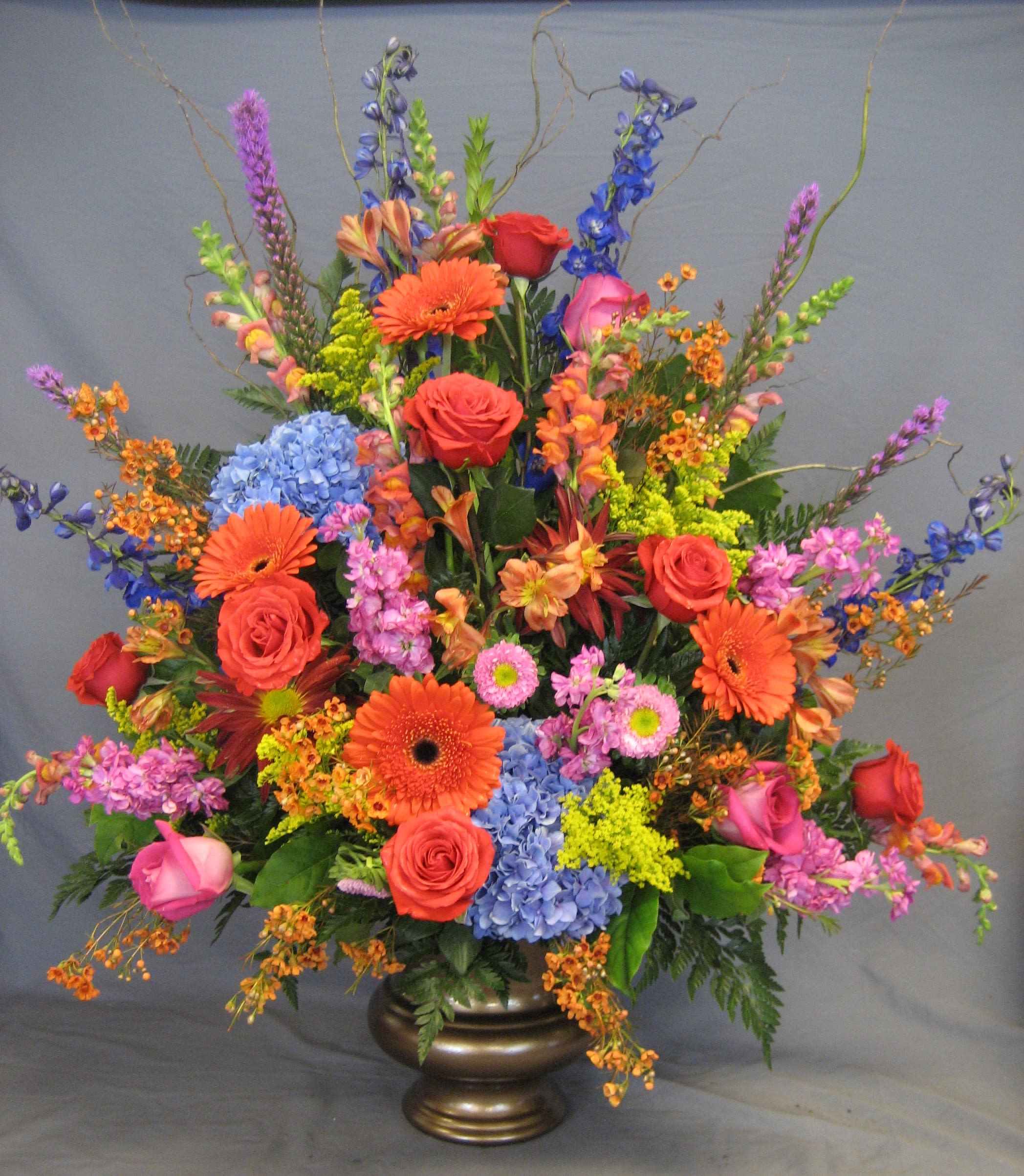 Bold Garden Mache M22 - Uplifting and heartwarming. This bold display of gerber daisies, hydrangea, roses, delphinium, asters and more are arranged expertly in a warm copper urn.