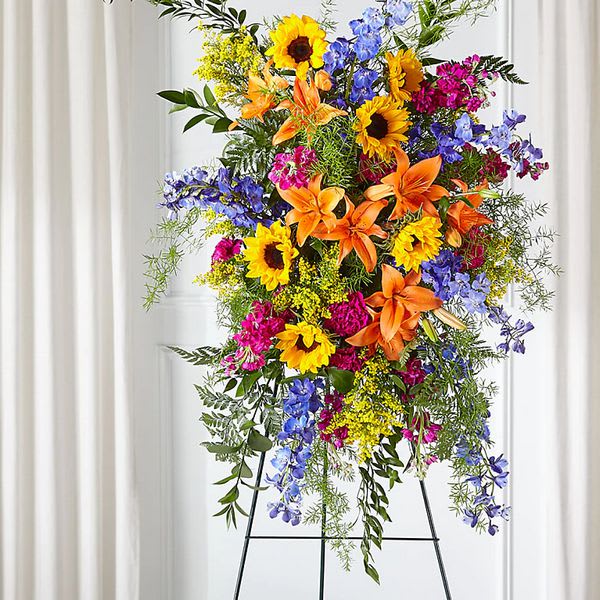 Sunlit Memories - Introducing our Sunlit Memories Standing Spray, a radiant and vibrant tribute to celebrate the life of your loved one. This stunning arrangement is crafted with a mix of fuchsia, deep blue, hot pink, and orange florals, creating a captivating display that reflects their vibrant personality and the joy they brought to those around them.  The Sunlit Memories Standing Spray is a celebration of life and a tribute to the colorful spirit of your loved one. The fuchsia blooms symbolize gratitude and grace, expressing your appreciation for the love and joy they brought into your life.  Deep blue florals add depth and a sense of tranquility to the arrangement. Blue is often associated with peace and serenity, reminding us of the comforting presence your loved one provided during their time with us.  The hot pink flowers represent love, passion, and admiration, reflecting the intense emotions and deep connection you shared with your departed loved one. These bold blooms serve as a reminder of the love and joy they brought into your life and the lives of others.  Adding a burst of energy and vibrancy to the arrangement are the orange florals. Orange is a color of enthusiasm and warmth, symbolizing the radiant and lively personality of your loved one. These bright blooms pay tribute to their zest for life and the positive impact they had on those around them.  The Sunlit Memories Standing Spray is a visual feast for the eyes and a fitting tribute to honor the life and legacy of your loved one. It is meticulously designed and showcased with care, ensuring that each flower and accent is arranged for maximum impact and beauty.  This standing spray serves as a striking and heartfelt tribute, capturing the essence of your loved one's radiant life. The combination of fuchsia, deep blue, hot pink, and orange florals creates a vibrant and dynamic arrangement that celebrates their spirit and brings comfort to those mourning their loss.  Choose our Sunlit Memories Standing Spray to honor and celebrate the vibrant life of your loved one. Let the bold and beautiful mix of colors serve as a reminder of the joy they brought into your life and the cherished memories that will forever be etched in your heart. 