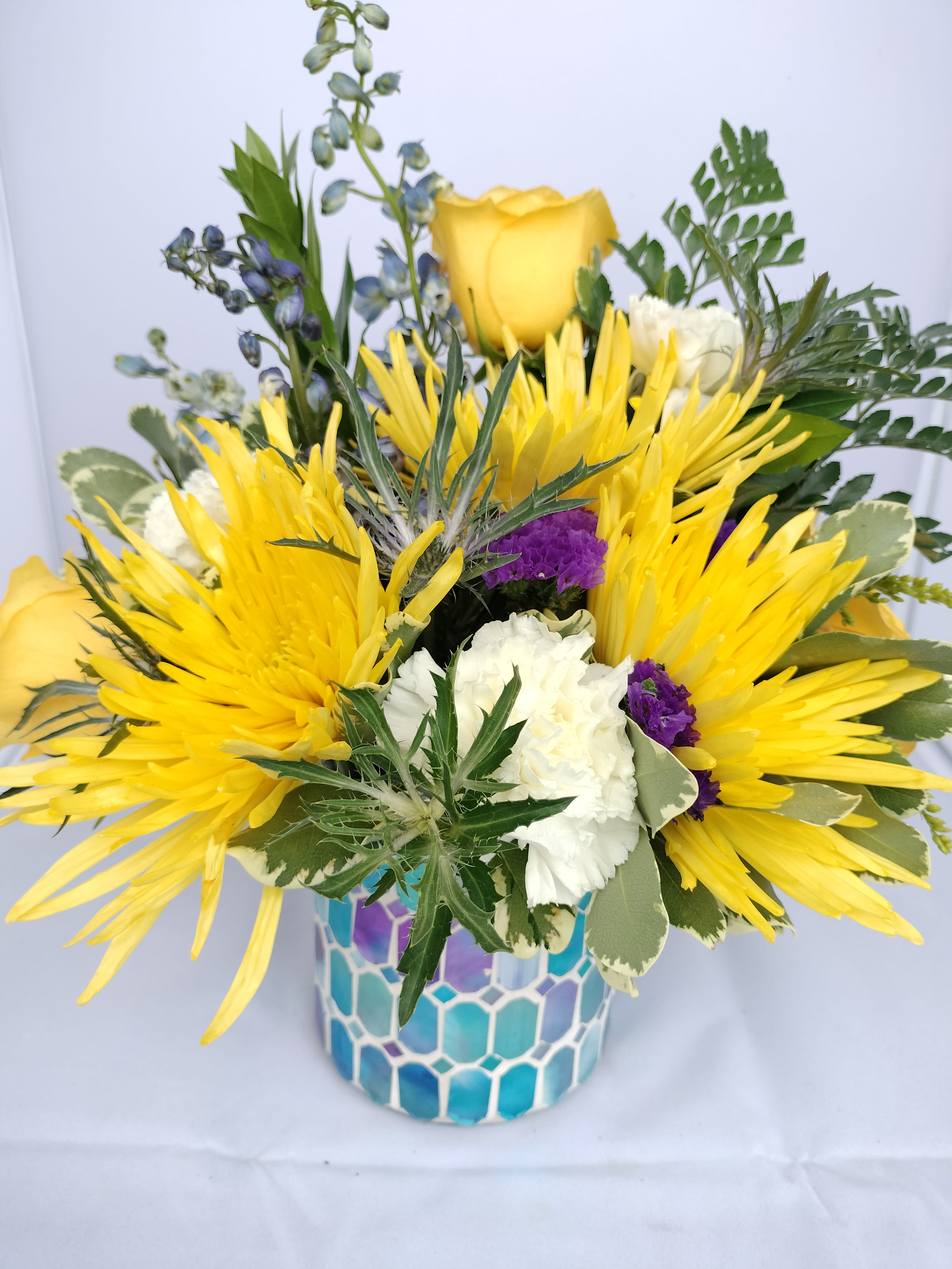 Dreams of the Shore - Dreams of a seaside escape should brighten their day and bring a smile to their face. This blue stained glass mosaic vase filled with blue delphinium, yellow spider mums, blue thistle, yellow roses and accents of purple statice will surely be a hit.