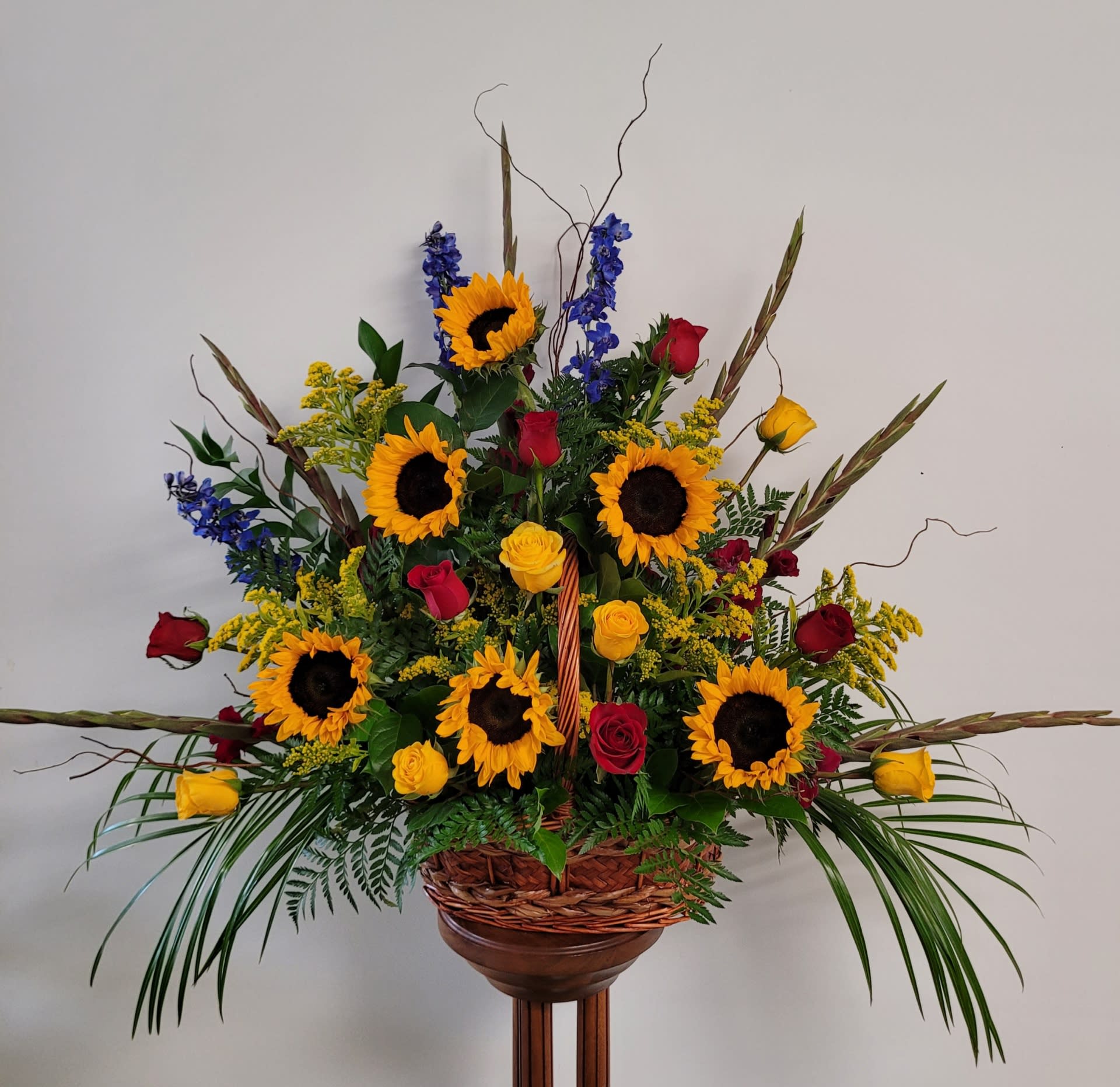Countryside Funeral Basket - Flowers such as sunflowers, gladiolus, delphinium, and roses in a wicker basket remind us of a country garden. Flowers may vary, due to availability.