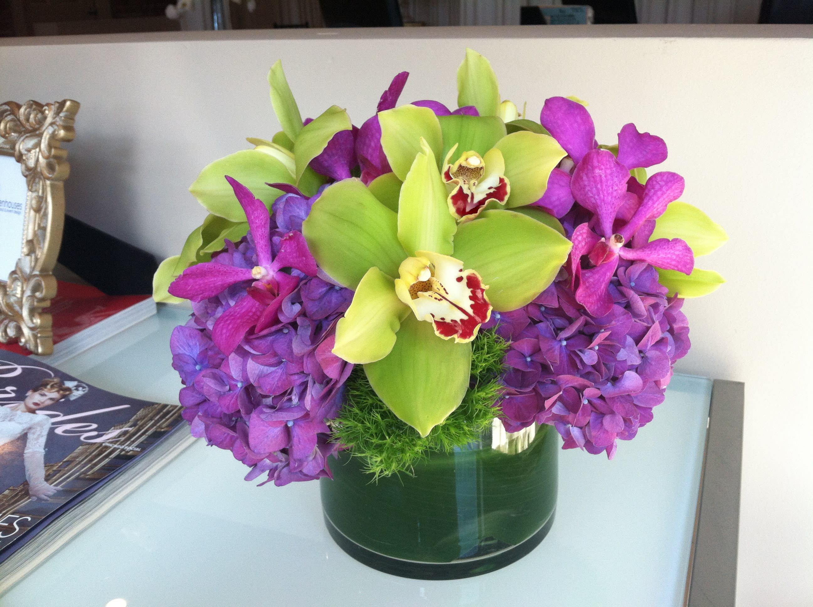 Orchid Overload Mini Orchid Overload Mini - Green cymbidium and magenta mokara orchid blooms with purple hydrangea and green dianthus in a 6x6" cylinder lined with a leaf wrap. Approximate arrangement size of 12x12".