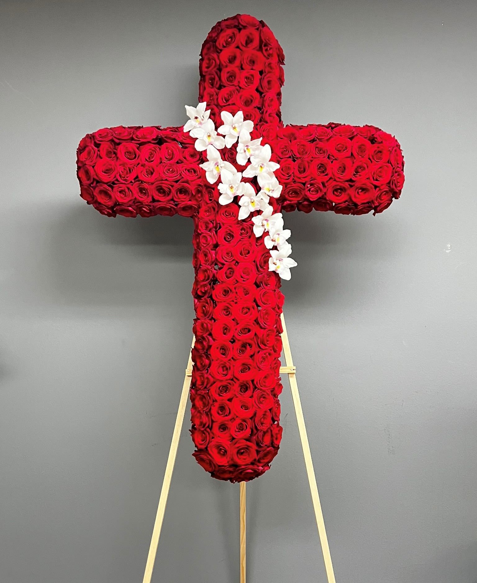 Red Cross with Orchids - This arrangement is a beautiful and symbolic tribute for a funeral service. It combines the elegance of red roses and the grace of cymbidium orchids to create a meaningful display. The arrangement is designed in the shape of a cross, representing faith, hope, and love. The cross is typically placed on an easel, allowing it to be prominently displayed during the service. The use of an easel ensures that the arrangement is visible from different angles, allowing everyone to appreciate its beauty. The choice of red roses symbolizes love, respect, and deep affection for the departed. Red roses are often associated with strong emotions and are commonly used in funeral arrangements to convey heartfelt condolences. In the center of the cross, there are cymbidium orchids. These orchids are known for their delicate and exotic beauty, symbolizing love, luxury, and strength. Their presence in the arrangement adds a touch of elegance and sophistication. Orchids are arranged with an angle signifying the ascension and transition from earthly life to the spiritual realm. It represents the journey of the departed soul towards eternal peace. Arrangement comes with black or white ribbon with professionally printed letters. Also, you can choose to attach a printed sympathy card. Just make a note during checkout.