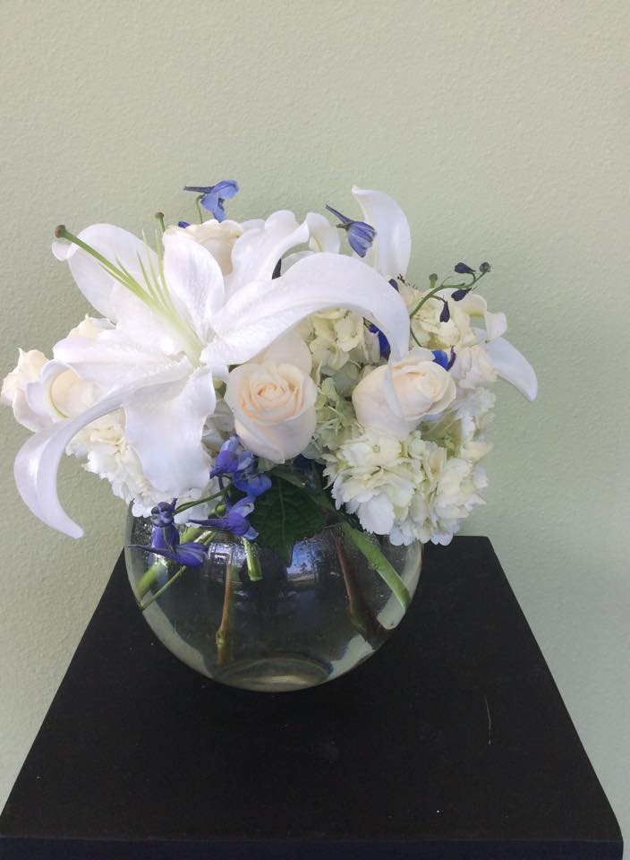 By The Seashore - A compact arrangement of white oriental lilies, white hydrangea, white roses, and blue delphinium in a circular glass bubble bowl. 