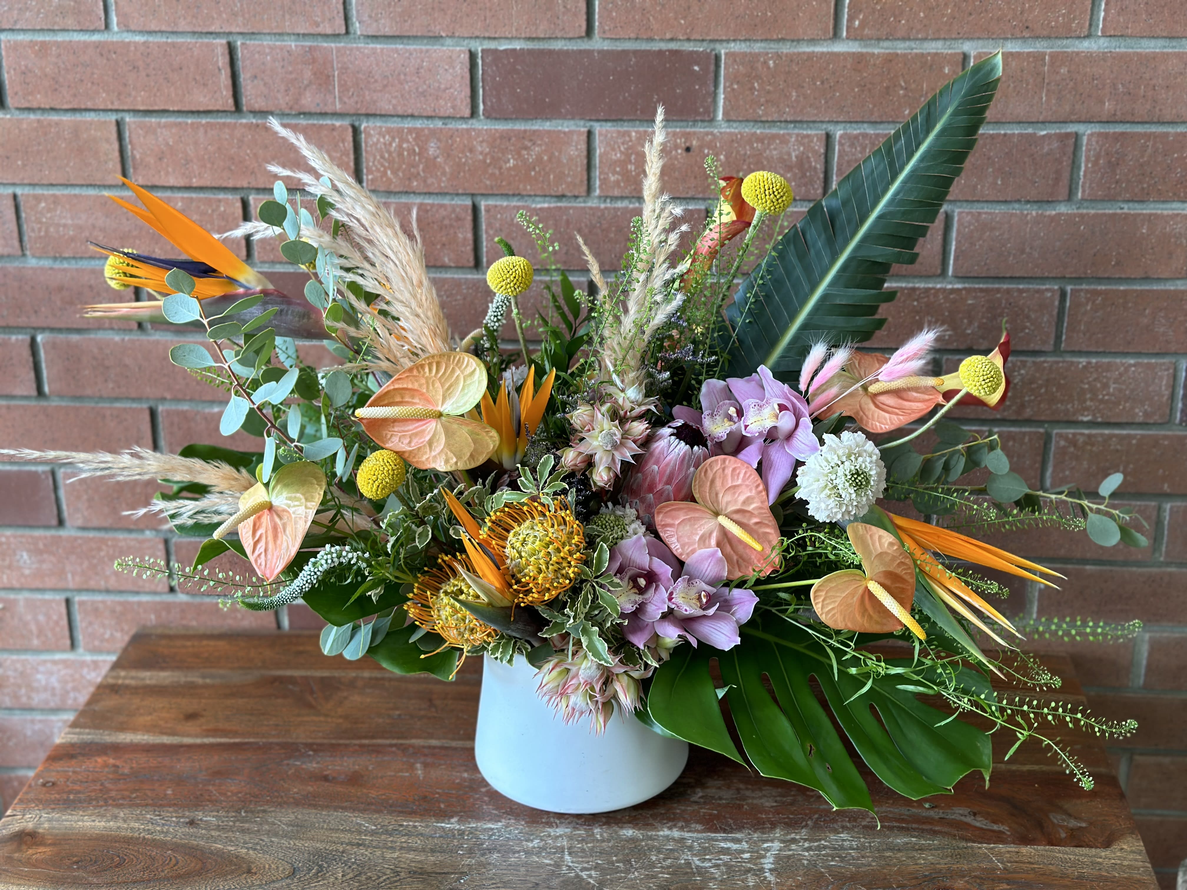 Coral Island Tropical Bouquet  - Modern, light, lush - a tropical arrangement filled with blush peach anthurium, fresh orchids, birds of paradise, monstera leafs, proteas and so much more. Designed in a modern white ceramic vase accented with pampas’s grass. 