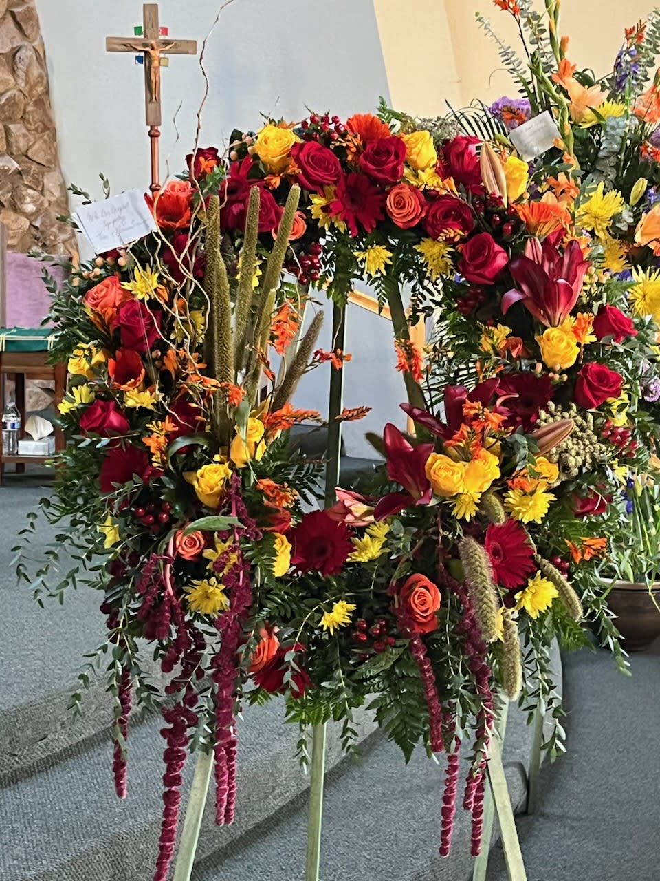 Large Fall Sympathy Wreath - A full garden selection of fall flowers arranged on a wreath form and delivered on a wooden stand. Pictured here are roses, gerbera, mums, hanging amaranthus, lilies, reeds and grasses. Seasonal substitutions will be made according to availability. Stands about 6ft tall and 3ft wide.