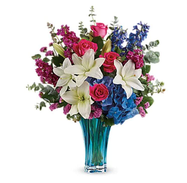 Teleflora's Ocean Dance Bouquet - As enchanting and elegant as the sea, this stunning blown glass vase is the most beautiful shade of azure and features a dramatic swirled shape. What a majestic way to present mom with a fabulous Mother's Day bouquet! Hot pink roses, white asiatic lilies, blue hydrangea, fuchsia stock, blue delphinium, and raspberry sinuata statice are arranged with lemon leaf and spiral eucalyptus. Delivered in a Heart's Pirouette vase.   Orientation : All-Around  All prices in USD ($)  Standard  T601-9A  Deluxe  T601-9B  Premium  T601-9C