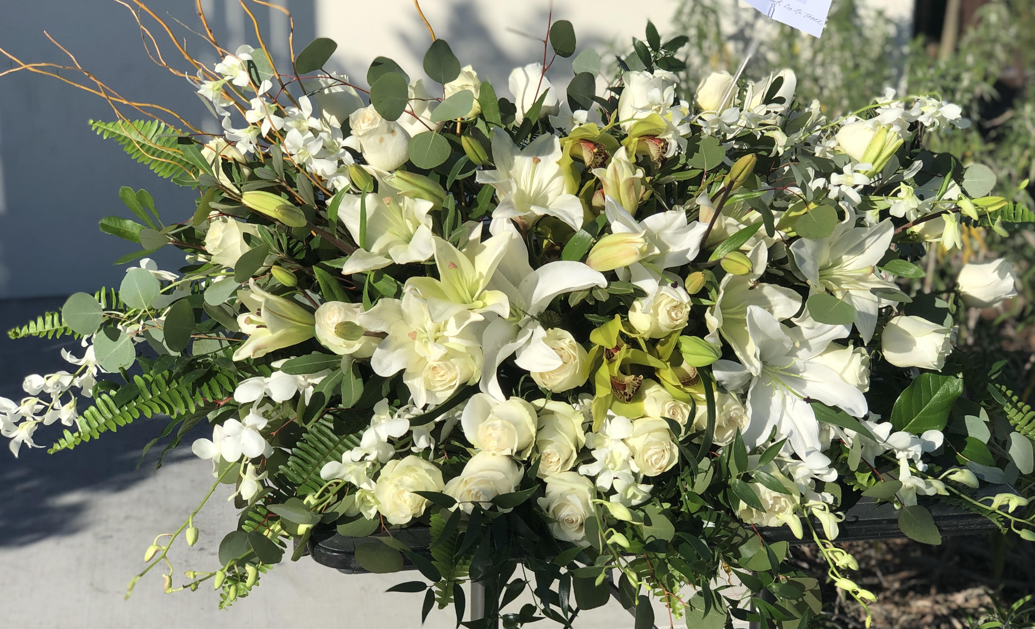 Grandiose Glorious Casket Spray - As serene as gently falling snow, this pure white spray of hydrangea, orchids, roses and lilies is a heartfelt symbol of peace and beauty--a symbol that will remain a guiding light to your loved ones for years to come. This stunning spray features white hydrangea, white cymbidium orchids, white dendrobium orchids, white roses, white spray roses, white oriental lilies, curly willow, green ivy, parvifolia eucalyptus, seeded eucalyptus, silver dollar eucalyptus, sword fern, and lemon leaf.