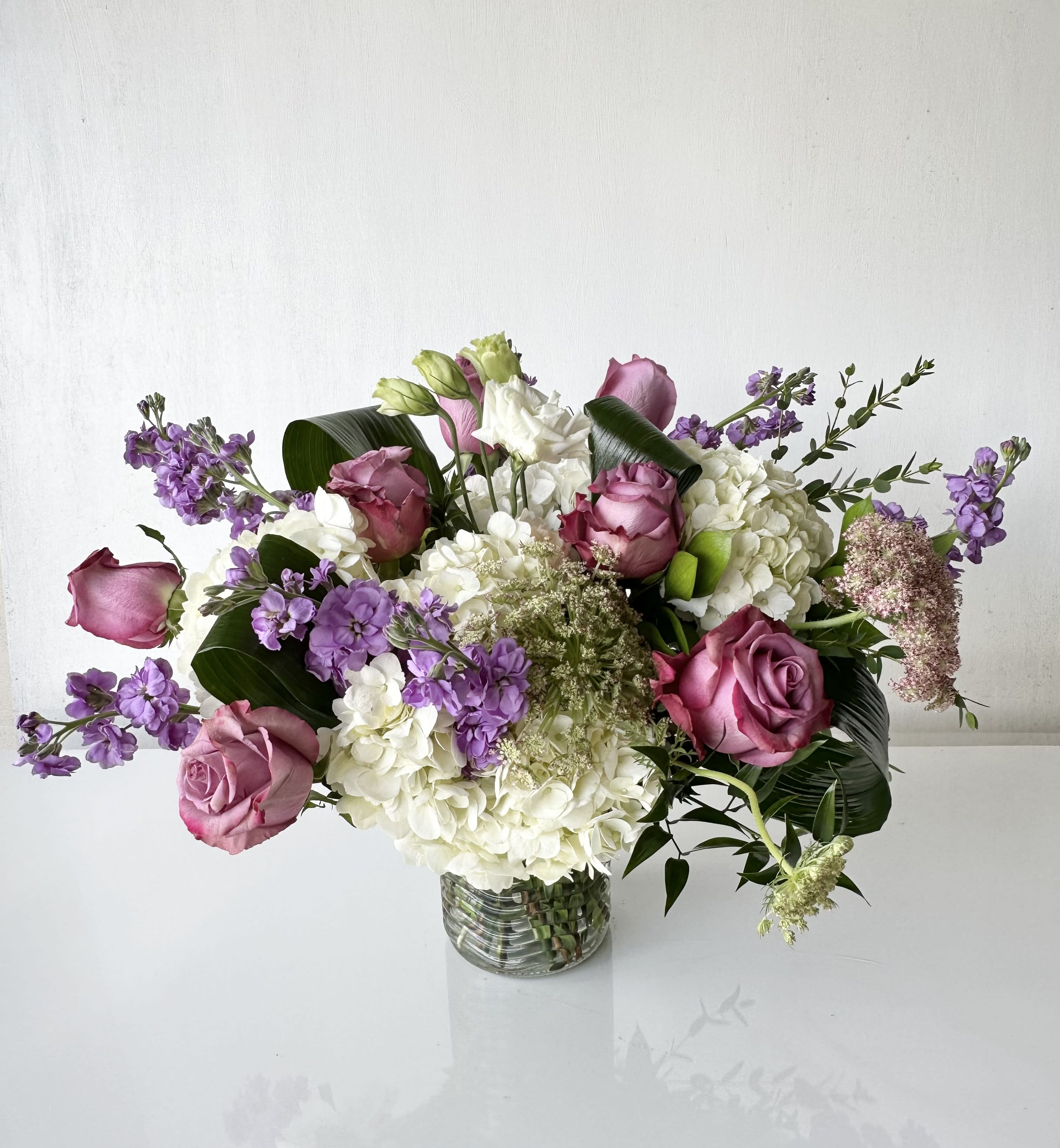 Polina - An arrangement of white hydrangeas, purple roses, lavender stock, and queen ann's lace with various greens in a glass vase.  Dimensions: Approximately 18&quot; H x 24&quot; W