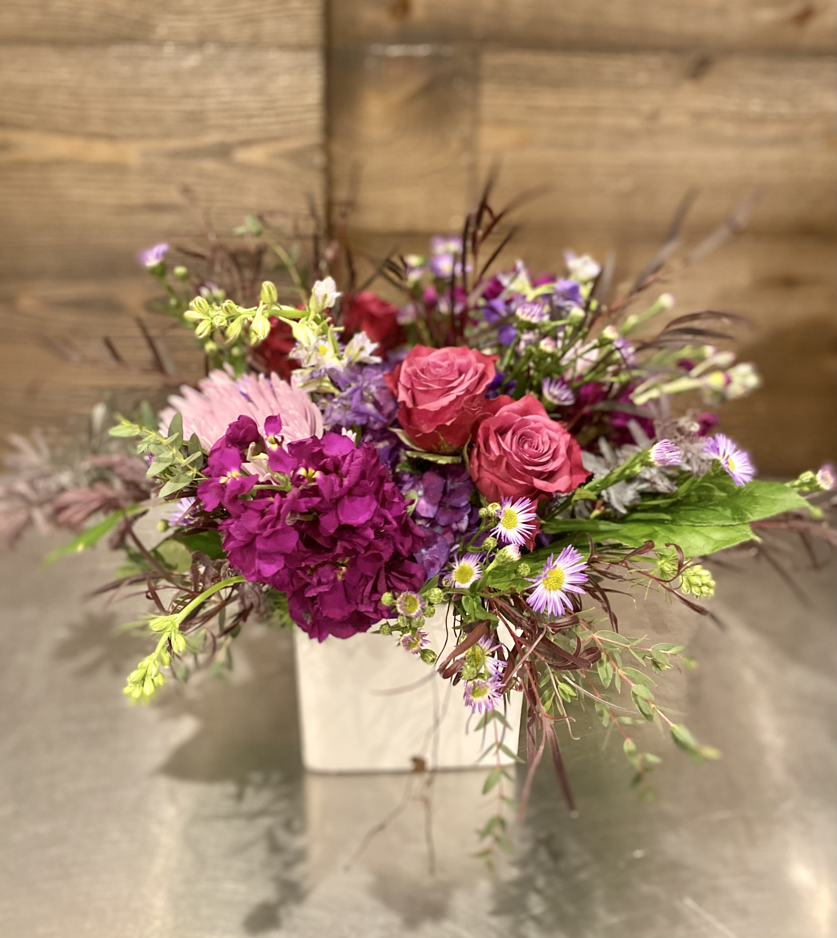 Purple Rain Purple Rain - Lush mix of purple and dark fuchsia flowers along with mixed greenery designed in a white ceramic square vase.