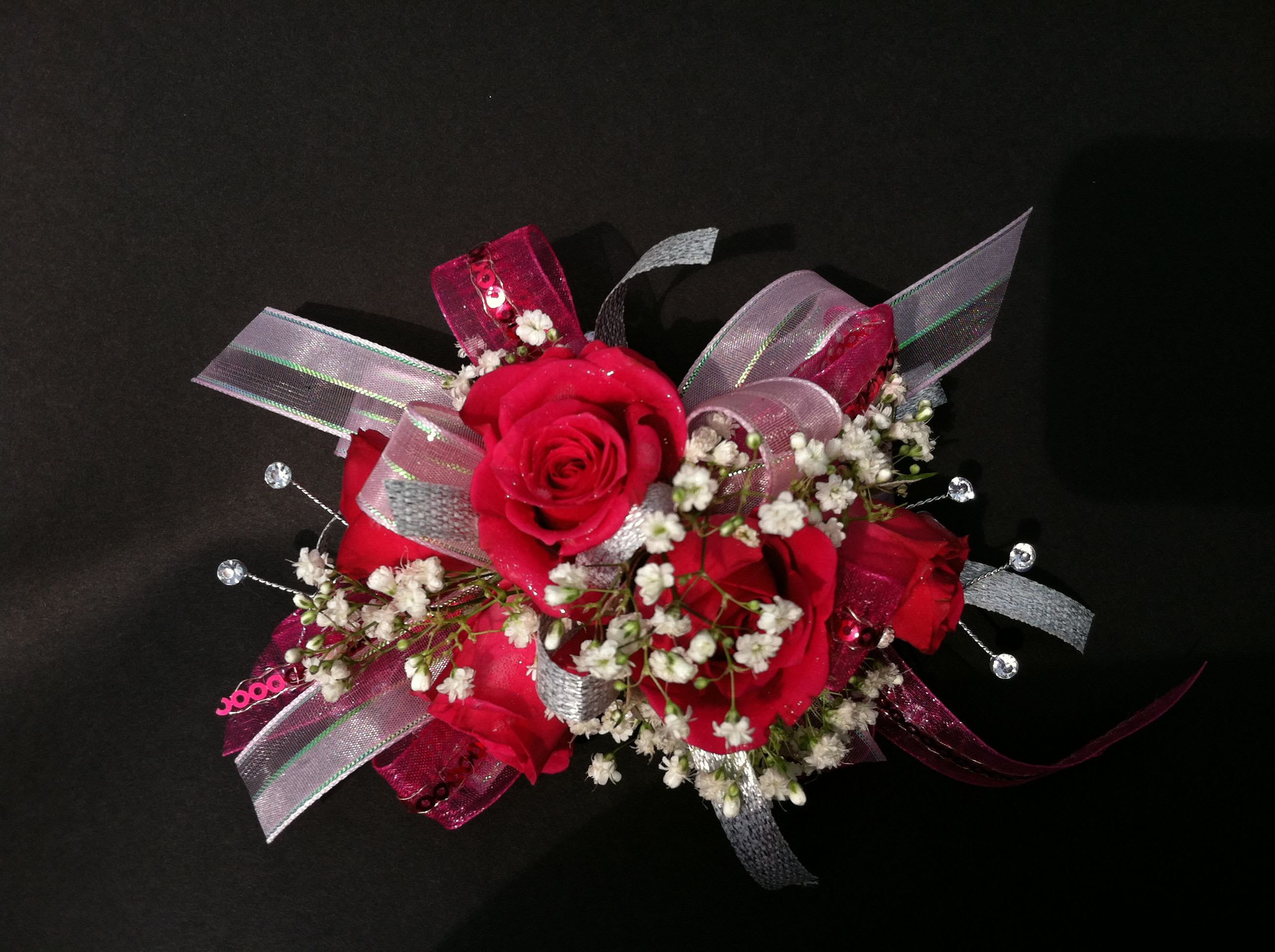 Red Rose Corsage Red Rose Corsage - Accent your formal wear with stunning flowers! Always in Season has an extensive collection of corsages, wristlets, and boutonnieres perfect for homecoming, prom, weddings, or any formal occasion. Accent your gown, tux, or any formal dress with our gorgeous floral designs! We customize each wrist corsage with matching accents of ribbon, diamonds, beads or accents. Please note in your order if you would like any other color of ribbon. Standard design includes bracelet, spray roses, accent filler flowers, and bow Deluxe design includes bracelet, spray roses, accent filler flowers, bow and either pearls or rhinestones. Premium design includes bracelet, spray roses, accent filler flowers, bow, rhinestones or pearls and bling design with upgraded bracelet.