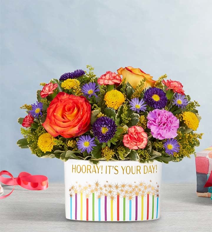 Hooray! It’S Your Day!™ Bouquet - Celebrate the special days, big and small, with our medley of bright blooms. Festively arranged in our keepsake “Hooray It’s Your Day” container, this happy surprise is sure to make the moment one they’ll remember. • All-around arrangement with bi-color yellow roses, purple Matsumoto asters and monte casino, yellow button poms, lavender carnations, orange mini carnations; accented with assorted greenery • Artistically designed in our “Hooray! It’s Your Day!” keepsake container adorned with colorful sparklers topped with metallic bursts of light