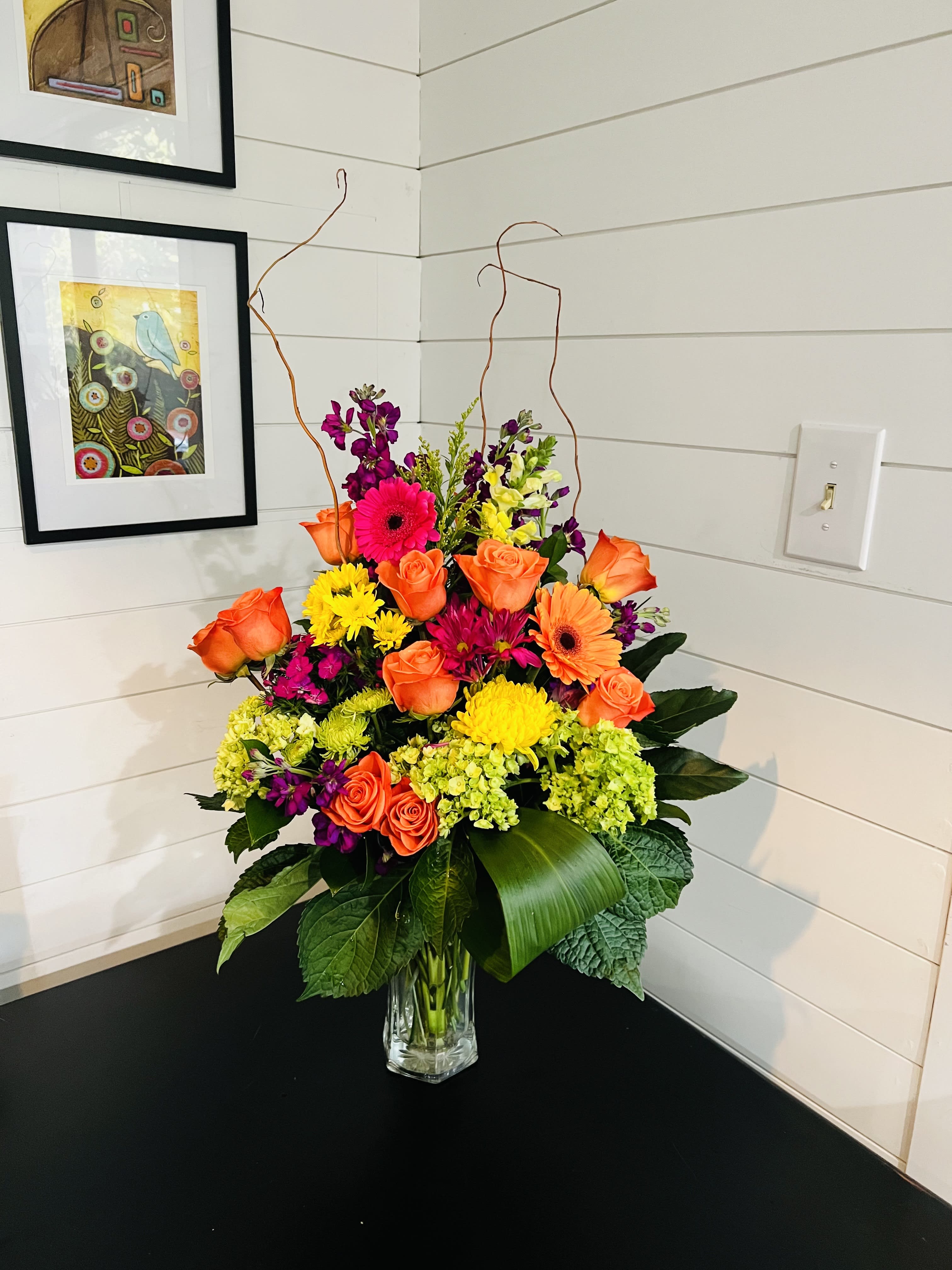 Friendship Blooms - A colorful, tall arrangement with a dozen orange roses, mini green hydrangea, purple stock, gerbera daisies, mixed fillers, curly willow and assorted greenery in a clear vase.