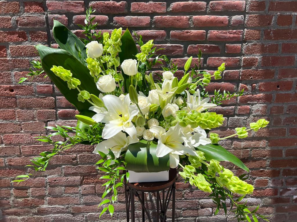 warm Tribute - composed of Ti Leafs, white lillies, white roses, bells of Ireland, Italian ruskus and white spray roses. In a modern white cube vase