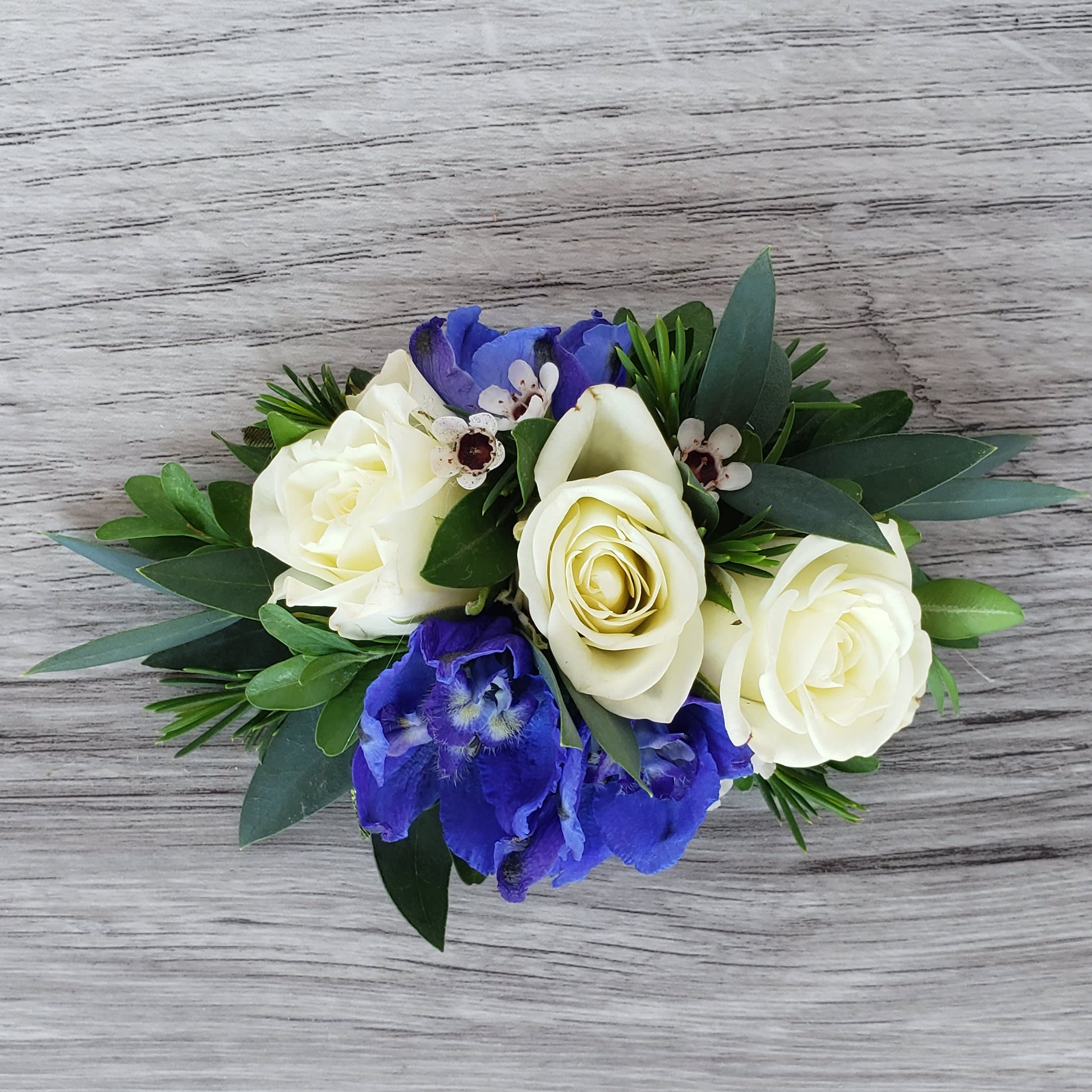 Blue and White Petite Corsage - This corsage is more petite in size and features white spray roses, blue delphinium, and complementary greens and accent flower. If you would like to exchange the colors of the flowers please let us know and we will be happy to accommodate. 