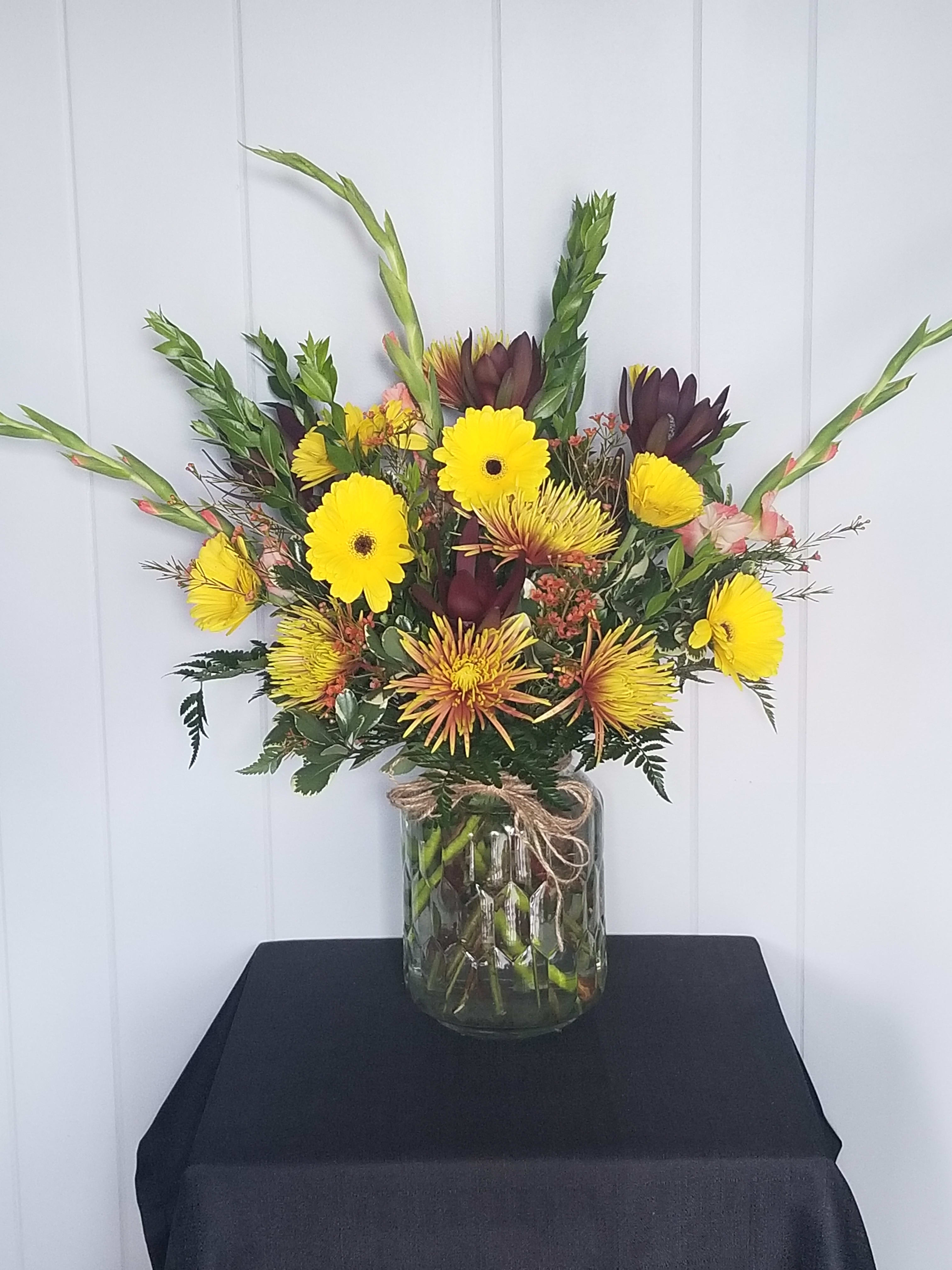 Fall in Love - This delightful arrangement of fall Gerbera Daisies, Mums, Gladiolus, surrounded by a fresh mixture of seasonal fillers and greens in a cute glass vase with a raffia bow. This arrangement looks great on anyone's table for any occasion! 