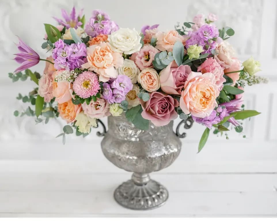 Positively perfect - Introducing our stunning modern floral arrangement, an exquisite combination of pink, peach, and lavender roses, spray roses, lisianthus, and more. This contemporary masterpiece is expertly arranged in a pedestal vase, adding an extra touch of elegance and sophistication to the design.  At the core of this arrangement are the captivating pink, peach, and lavender roses that steal the spotlight. Carefully selected for their pristine quality and captivating hues, these roses symbolize love, beauty, and grace. The harmonious blend of soft and delicate colors creates a romantic and dreamy ambiance, instantly captivating any onlooker.  Complementing the roses are the delicate spray roses. These petite blooms add a whimsical touch and bring a sense of playfulness to the arrangement. Their clusters of tiny blossoms in various shades of pink, peach, and lavender create a visually stunning and textured display. The spray roses elevate the arrangement's visual interest, making it truly unique.  Adding an air of refinement and elegance are the lisianthus blooms. With their delicate petals and graceful presence, the lisianthus flowers perfectly complement the roses and spray roses. Available in shades of pink, peach, and lavender, the lisianthus brings a touch of sophistication to the overall arrangement, creating a harmonious blend of floral beauty.  To accentuate the natural beauty of these blooms, fresh eucalyptus leaves are thoughtfully intertwined throughout the arrangement. The lush green foliage adds an element of texture, depth, and dimension. In addition, the eucalyptus leaves lend a subtle and refreshing aroma that enhances the overall sensory experience.  All of these exquisite blooms, including the roses, spray roses, lisianthus, and eucalyptus, are meticulously arranged in a pedestal vase. The sleek and modern design of the pedestal vase elevates the arrangement, making it a true centerpiece that draws attention. The transparency of the vase allows the beauty of each bloom to shine, creating a visually captivating statement.  This modern floral arrangement in a pedestal vase is perfect for those who appreciate contemporary sophistication. Whether it's for home decor, a special gift, or an elegant centerpiece for a memorable event, this arrangement is sure to make a lasting impression.  Embrace the beauty of modernity with this enchanting floral arrangement. Let the combination of pink, peach, and lavender roses, spray roses, lisianthus, and eucalyptus, arranged in a pedestal vase, bring elegance and sophistication to any space or occasion. 