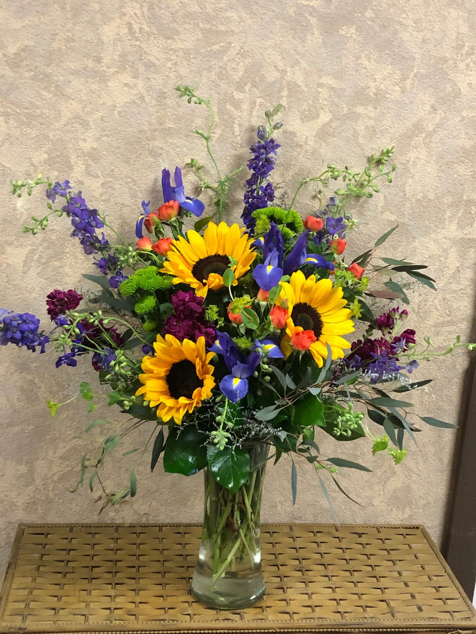 Summer Iris - Recipe: Purple Larkspur, Sunflowers, Purple Iris, Orange Spray Roses, Burgundy Stock, Green buttons, and greens in a tall vase. Availability: All year round Substitute Available: Yes Design View: Symmetric Front Facing View Photo shown: Regular