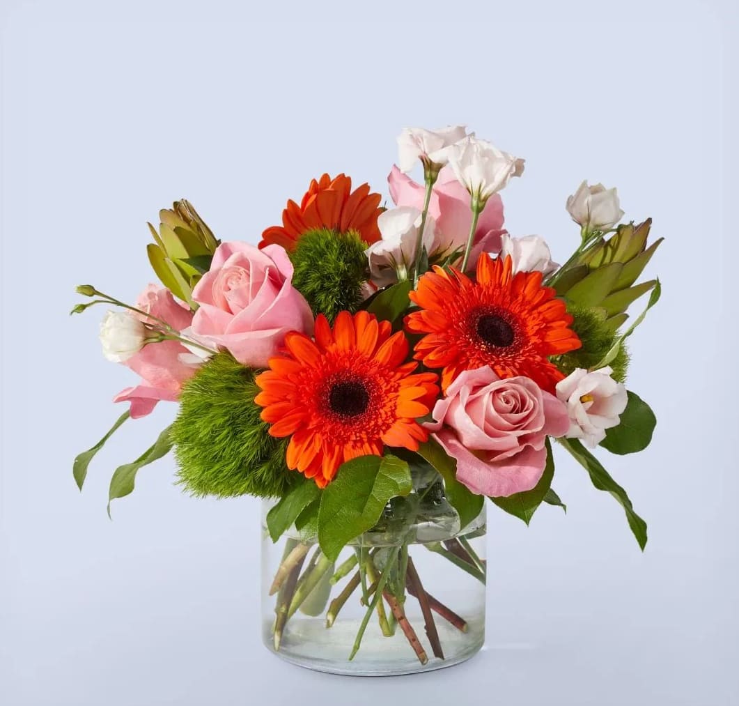 Feels Like Home - Experience the heartwarming embrace of &quot;Feels Like Home,&quot; a captivating arrangement that effortlessly brings comfort, warmth, and a sense of belonging. This beautiful bouquet showcases a stunning combination of pink roses, orange gerbera daisies, and green balls, artistically arranged in a clear glass vase.  At the core of this arrangement are the enchanting pink roses, representing love, gratitude, and admiration. Their exquisite blooms are a testament to the beauty and joy found within the comforts of home. Their delicate petals and soft fragrance evoke a sense of serenity and tenderness, making them the perfect choice for conveying heartfelt emotions.  Complementing the roses are the vibrant orange gerbera daisies, radiating happiness, joy, and positive energy. Their cheerful and vivid petals add a burst of color and playfulness to the arrangement, while their vibrant presence captures the essence of the familiar and welcoming nature of home.