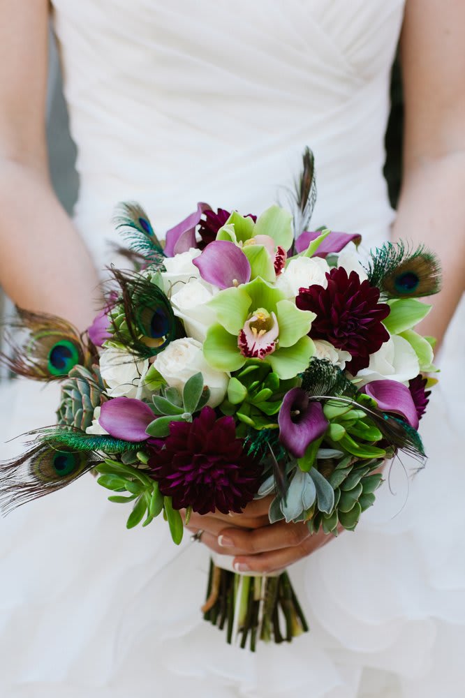 Divine Bridal Bouquet with Peacock Feathers - This unique bouquet radiates with peacock feathers, succulents, hot pink calla lilies, ruby red dahlias, white roses, and green orchids tied with ribbon as a hand held bouquet. Availability: All year round Substitution available: Yes  Must be ordered at least 5 days in advance.