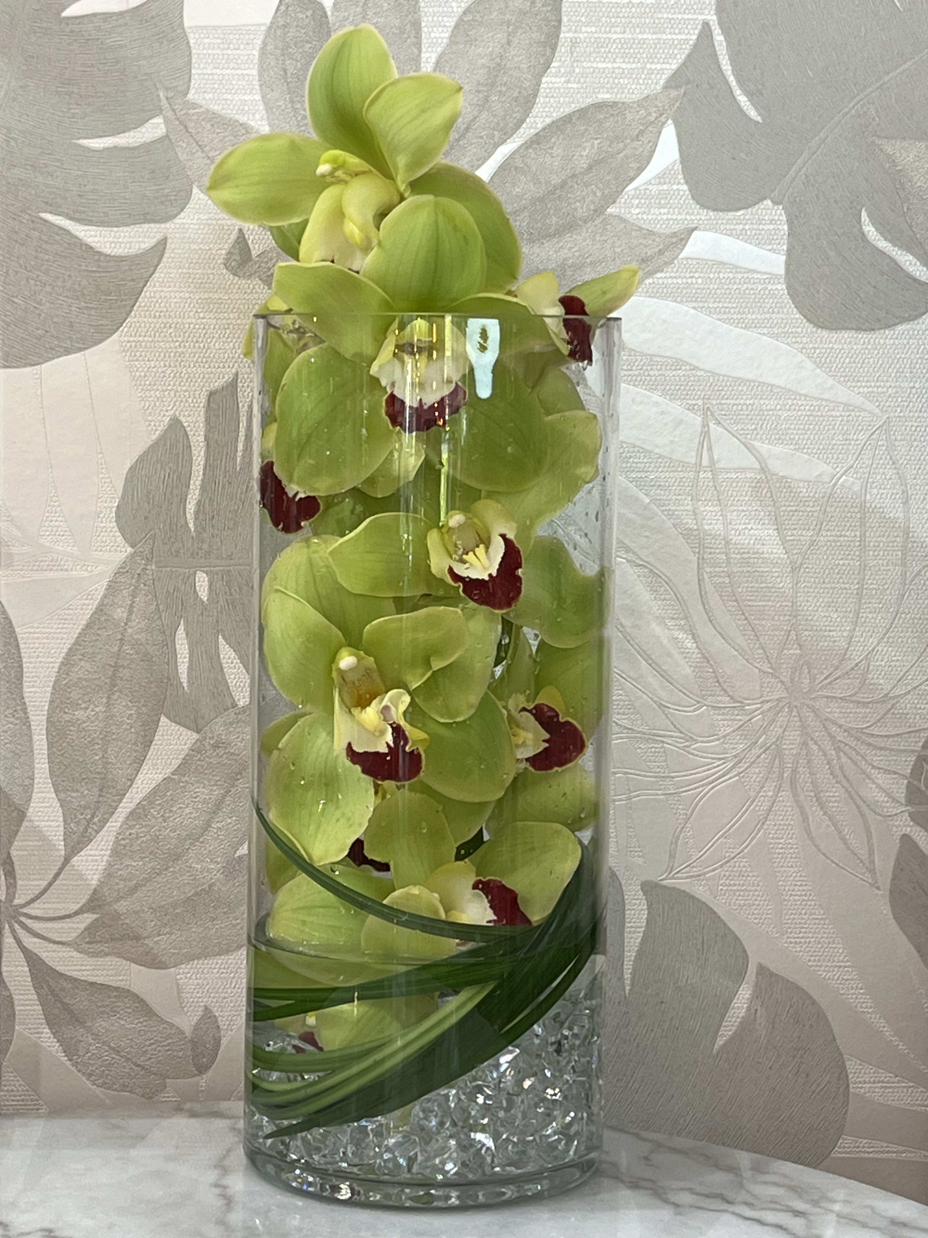 Simple Elegance - An emotionally soothing and unique design.  A full stem of Thailand or New Zealand cymbidium orchids in bloom, submerged in water anchored by iridescent glass stones.  The orchid is incased in a 16” tall glass cylinder vase.  Best for large offices, lobbies and homes.    