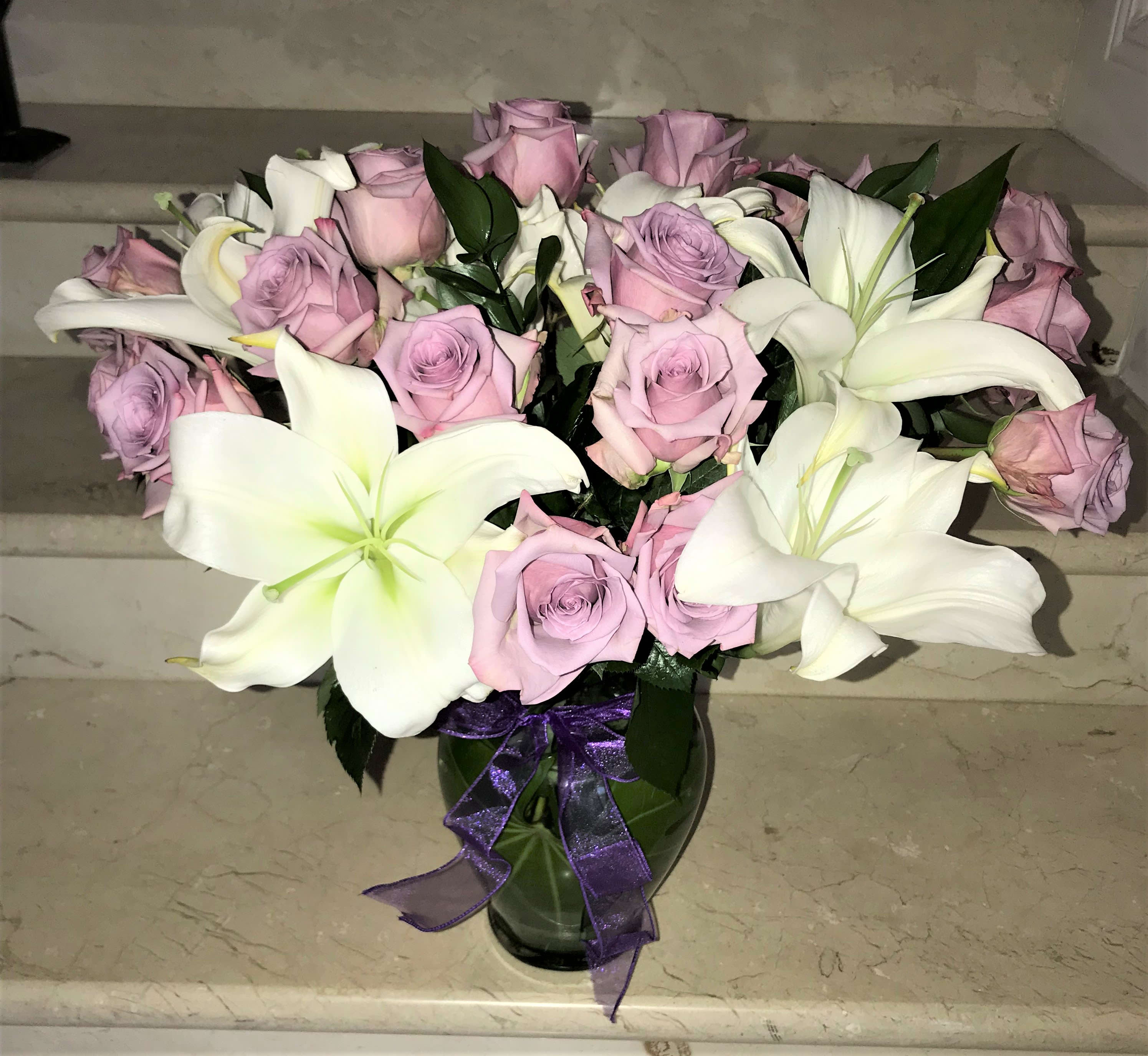 Lavender Roses and White Lilies - As Shown: has 18Lavender roses with white Lilies arranged with greens in a vase. Deluxe Upgrade 24 roses with white lilies with greens in a vase. 