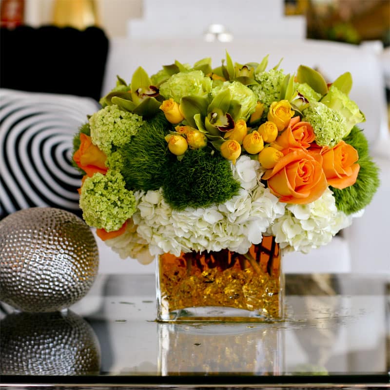 Liquid Gold - Green and orange roses, green and white hydrangeas, and pale green cymbidium orchids in a glass vase with golden water. 