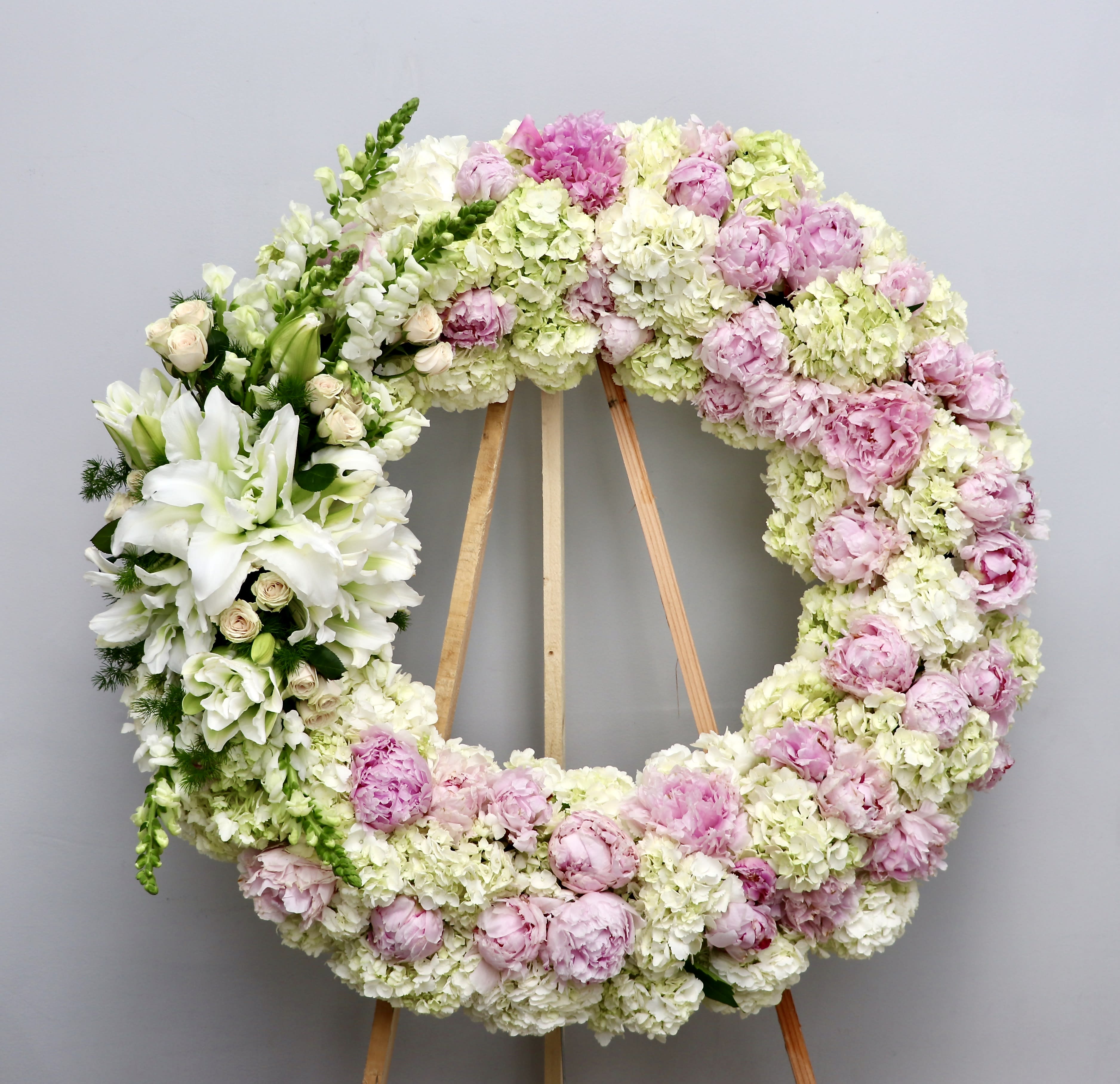 Peony and Hydrangea Wreath - With a mix of blush peony (garden roses when not in season) and hydrangeas, this sympathy wreath also contains snapdragons and lilies.   We include easel, printed banner and delivery (some fees may apply).  Standard size is 30'', deluxe is 36'', and premium is 42''.