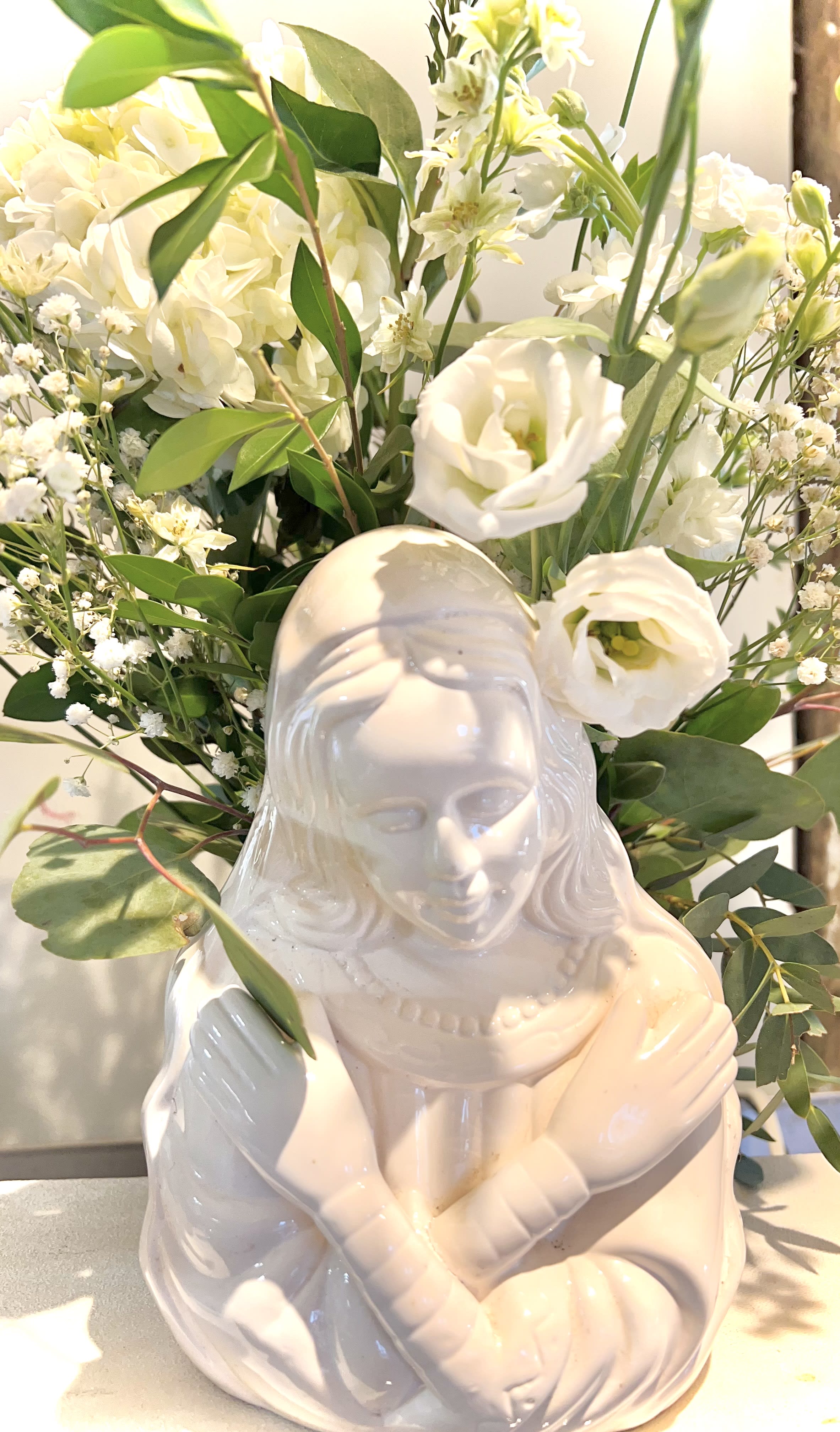 Blessed Morher bouquet Blessed Morher bouquet - A mixed arrangement of all white seasonal blooms (may vary depending on time of year) Arranged in a ceramic Blessed Mother vase