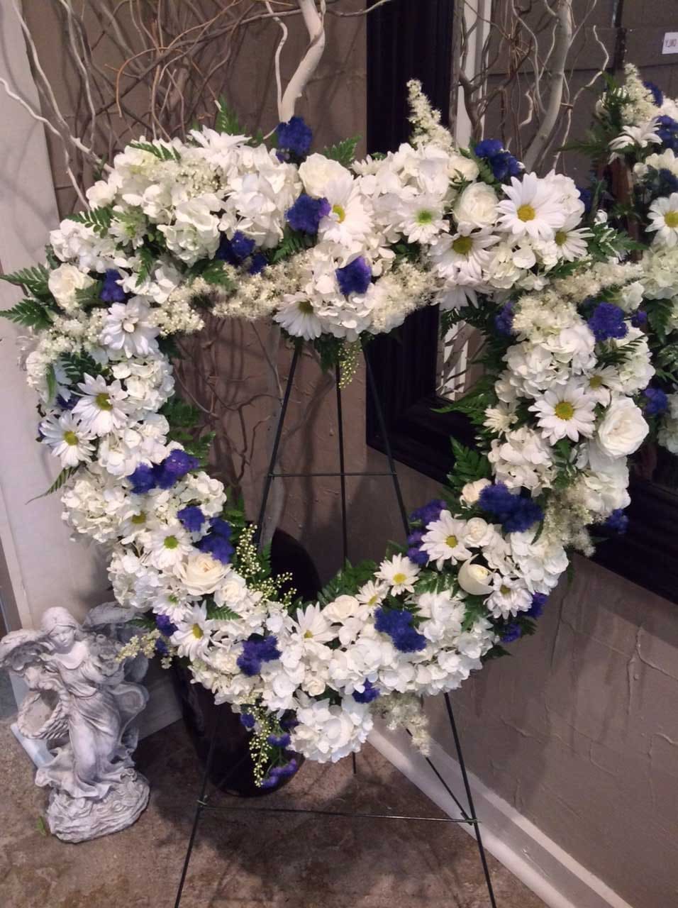 Missing You Large Standing Heart  - This delicate heart-shaped wreath is made with a lovingly handpicked selection of blue, cream, and white flowers. Perfect for a memorial service, this wreath evokes a sense of peace and remembrance with its intricate design. The combination of flowers create a serene and calming feel, allowing the memory of your loved one to be celebrated in a meaningful way.