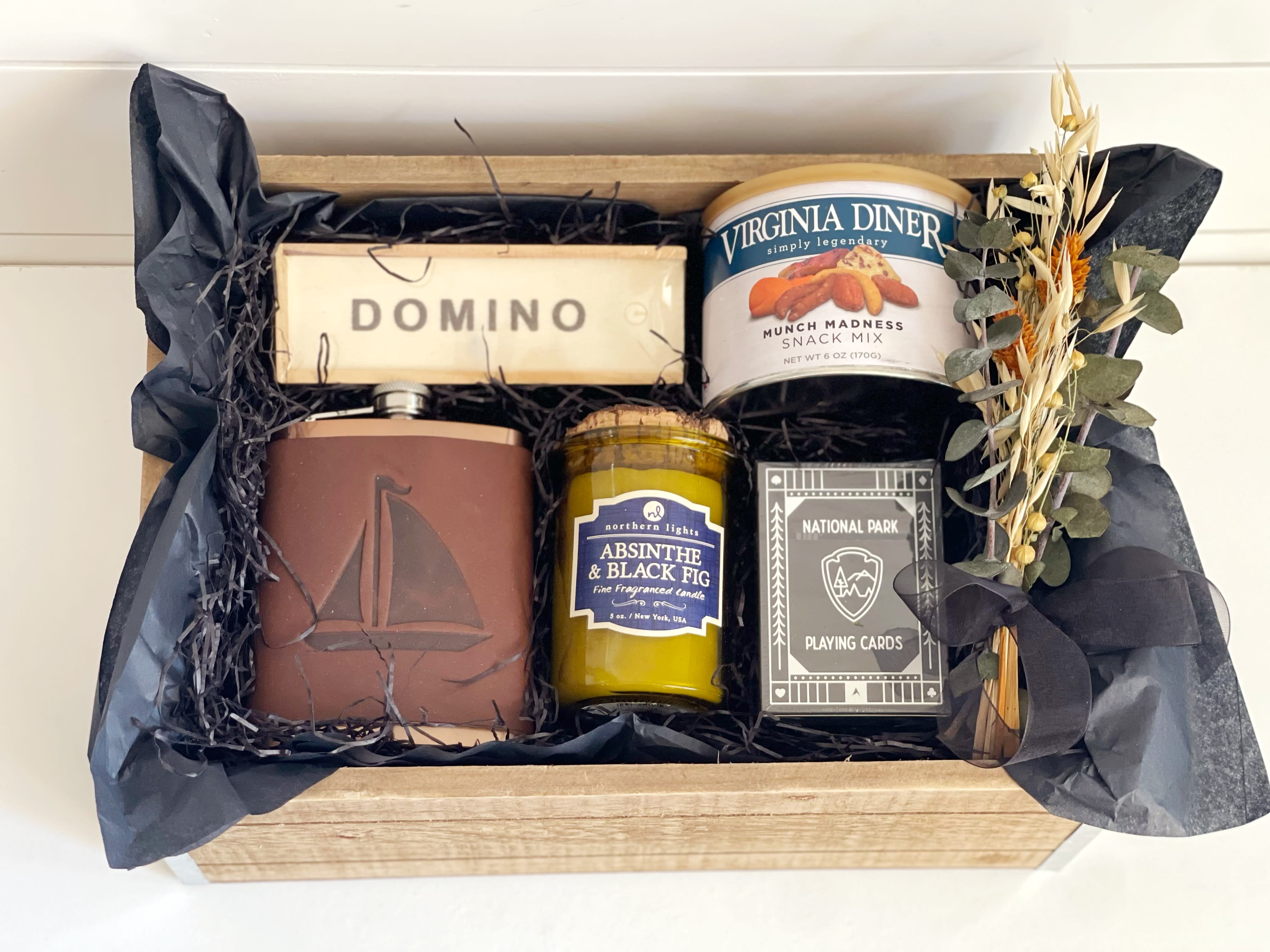 Man Cave Upgraded! - Show him your thinking of him in ways he’ll appreciate with this masculine curated gift basket. -National Park playing cards -wooden domino set Virginia Diner snack mix -Northern Lights Candle, this line combines fragrances and the wit of your favorite bartender. Fragrance notes: black fig infused with a woodsy blend of absinthe, black currant and patchouli. -Sailboat Sailing Leather Wrapped Flask hand made by Jimmyrockit. This is an 8 ounce stainless steel flask wrapped in a leather sleeve and hand stitched on the back. The flask has an attached hinged top and the leather is earthy, a perfect match.   