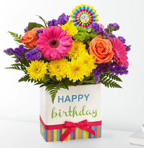 Birthday Brights Bouquet - The Birthday Brights Bouquet is a true celebration of color and life to surprise and delight your special recipient on their big day! Hot pink gerbera daisies and orange roses take center stage surrounded by purple statice, yellow cushion poms, green button poms, and lush greens to create party perfect birthday display. Presented in a modern rectangular ceramic vase with colorful striping at the bottom, &quot;Happy Birthday&quot; lettering at the top, and a bright pink bow at the center, this unforgettable fresh flower arrangement is then accented with a striped happy birthday pick to create a fun and festive gift.
