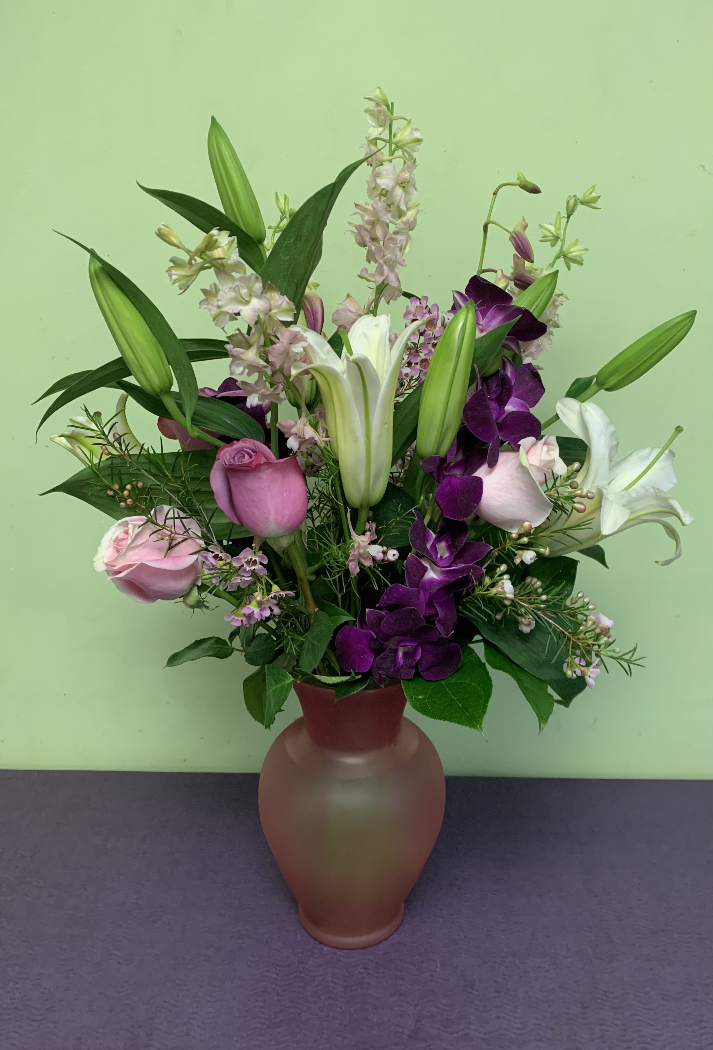 Purple Heart  - Melt her heart today with this stunning floral arrangement that is truly breath taking. If you want something that's a little more classy and elegant, then this arrangement is perfect for any one who has your heart! The arrangement arrives in either a pink or a purple vase. It also includes fragrant Hybrid Lilies and tropical orchids. There are Roses, Larkspur and Wax flower  added to this incredible arrangement.