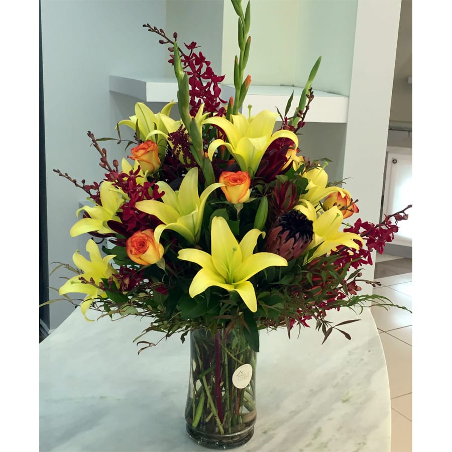 The Firecracker Bouquet - A very unique fiery mix of texture and color, this bouquet is not your traditional autumnal arrangement but perfect for the year round flower lover. This premium assortment features exotic Red Elephant Orchids, Proteas, Lilies, Roses and Gladiolus - all professionally hand arranged by our designers in a clear glass vase. Product is pictured in full / round style design which is a Deluxe version. Standard size is a one-sided presentation and measures approximately 18in (W) x 29in (H). Deluxe and Premium versions are larger.  Standard - One-Sided Presentation (Stem Count) - 4 Lilies, 3 Gladiolus, 5 Roses, 3 Red Elephant Orchids, 4 Safari Sunset Proteas, 1 Pink Ice Protea and Fresh Garden Greens - Designer Glass Vase  Deluxe - Full / Round Style Design (Stem Count) - 5 Lilies, 3 Gladiolus, 9 Roses, 4 Red Elephant Orchids, 5 Safari Sunset Proteas, 2 Pink Ice Protea and Fresh Garden Greens - Designer Glass Vase  Premium - Jumbo / Round Style Design (Stem Count) - 6 Lilies, 4 Gladiolus, 12 Roses, 5 Red Elephant Orchids, 6 Safari Sunset Proteas, 3 Pink Ice Protea and Fresh Garden Greens - Designer Glass Vase  Please Note: Upon delivery most lilies will be in bud form to ensure freshness and extended flower's life span. As with any lily, the stamens should be plucked off as soon as they present themselves from within the flower. This will enhance the life of the flower and prevent staining from the pollen.  Care Tips: Place your bouquet in a cool location. Don't put the arrangement in direct sunlight, near heating or cooling vents, in drafty places, directly under ceiling fans, or on top of televisions or radiators. Check water level daily, keep the vase full with clean water. Change water every 2-3 days and apply a sharp fresh cut to the stems. This process will ensure extended flower's life span.