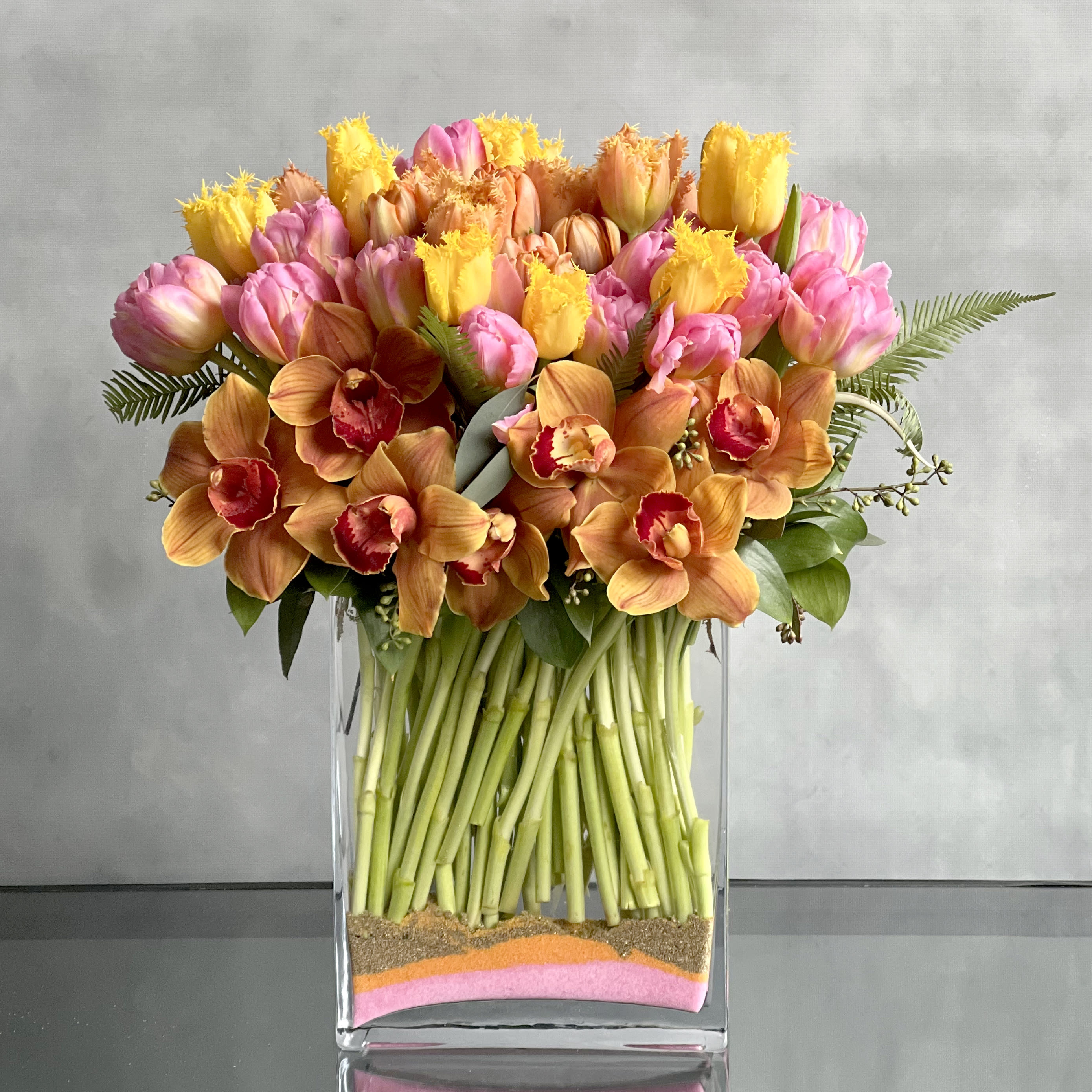 L'Orange!  - A modern twist to a classic flower! Various shades of colorful Tulips arranged within a rectangular vase, which is Accented with Orange Orchids. Vibrant and fresh just in time for the warming seasons. 