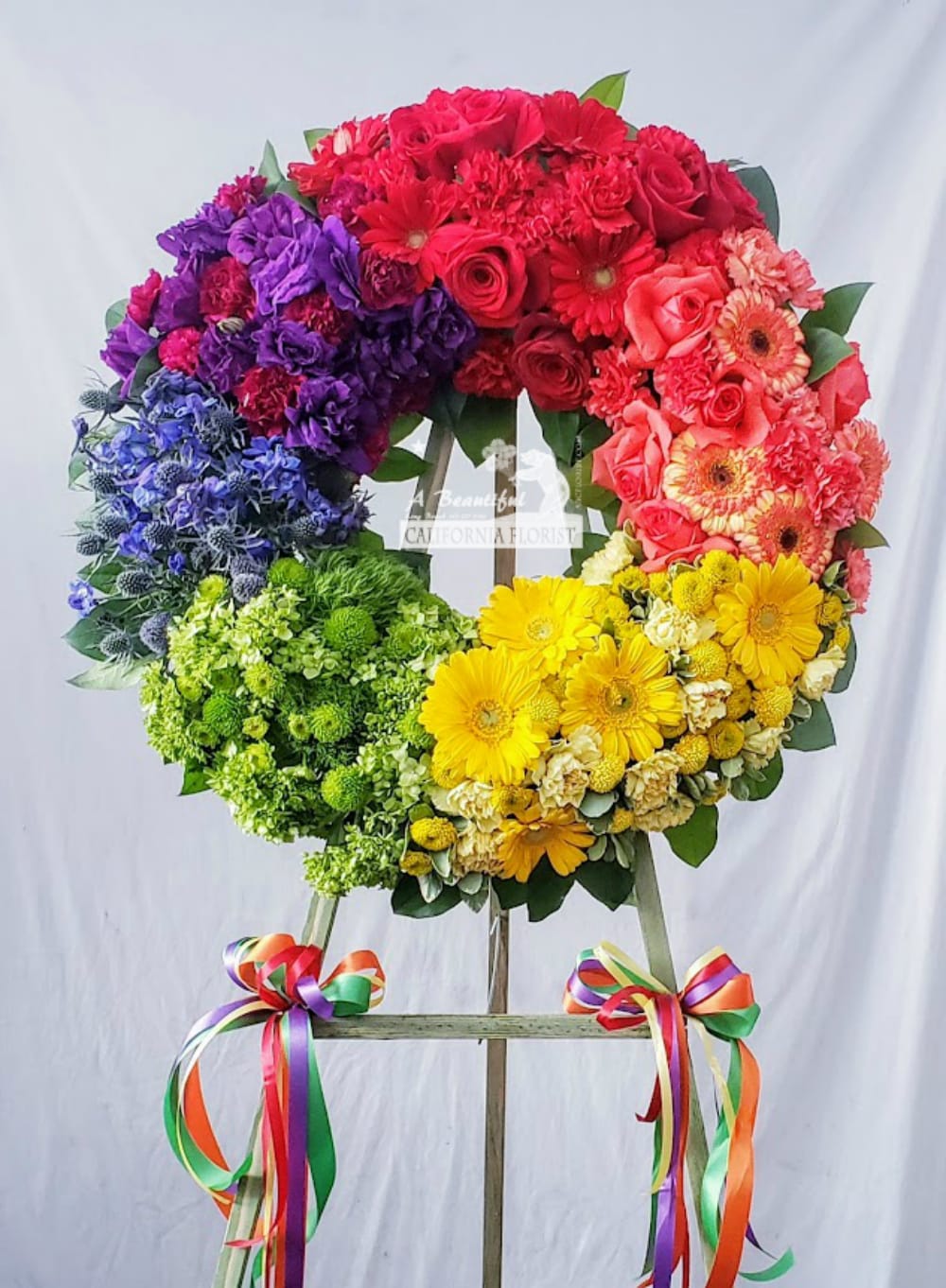 Rainbow of Memories Wreath  - &quot;Do not mourn for me in black, but paint me in a rainbow of color.&quot;  Rainbow of Memories, friends, family, laughter and love&quot; Beautiful Rainbow of Memories Wreath in an array of floral blooms in the colors of a rainbow, comes displayed on  wooden easel. Approximate wreath finished size 22&quot; diameter.