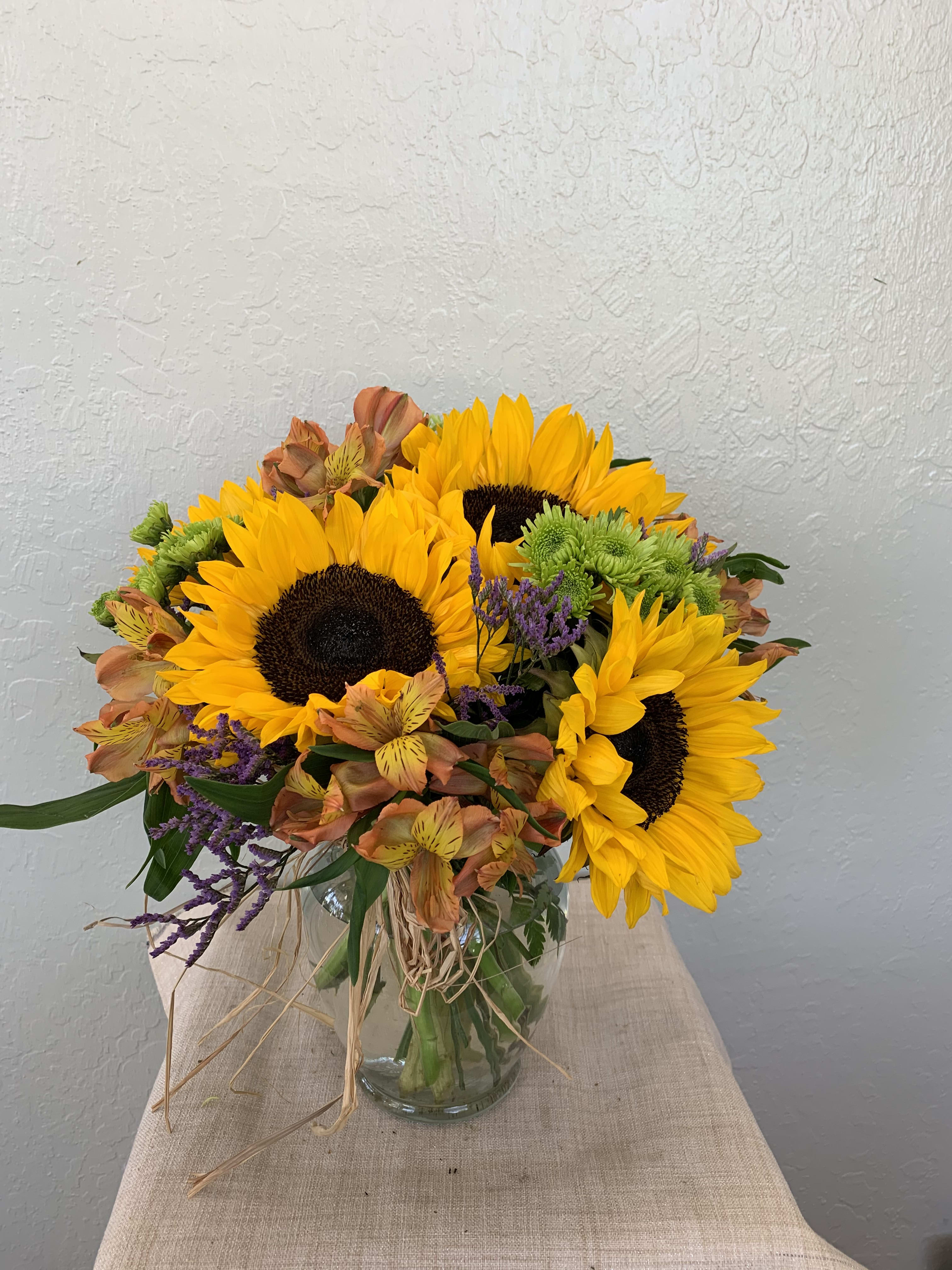 Sunflower Harvest Sunflower Harvest - A beautiful sunflower harvest filled with sunflowers, alstroemerias, pompoms, and seasonal filling greens in a clear glass vase wrapped in raffia. Approximately 14"H x 14"W Orientation: All-Around